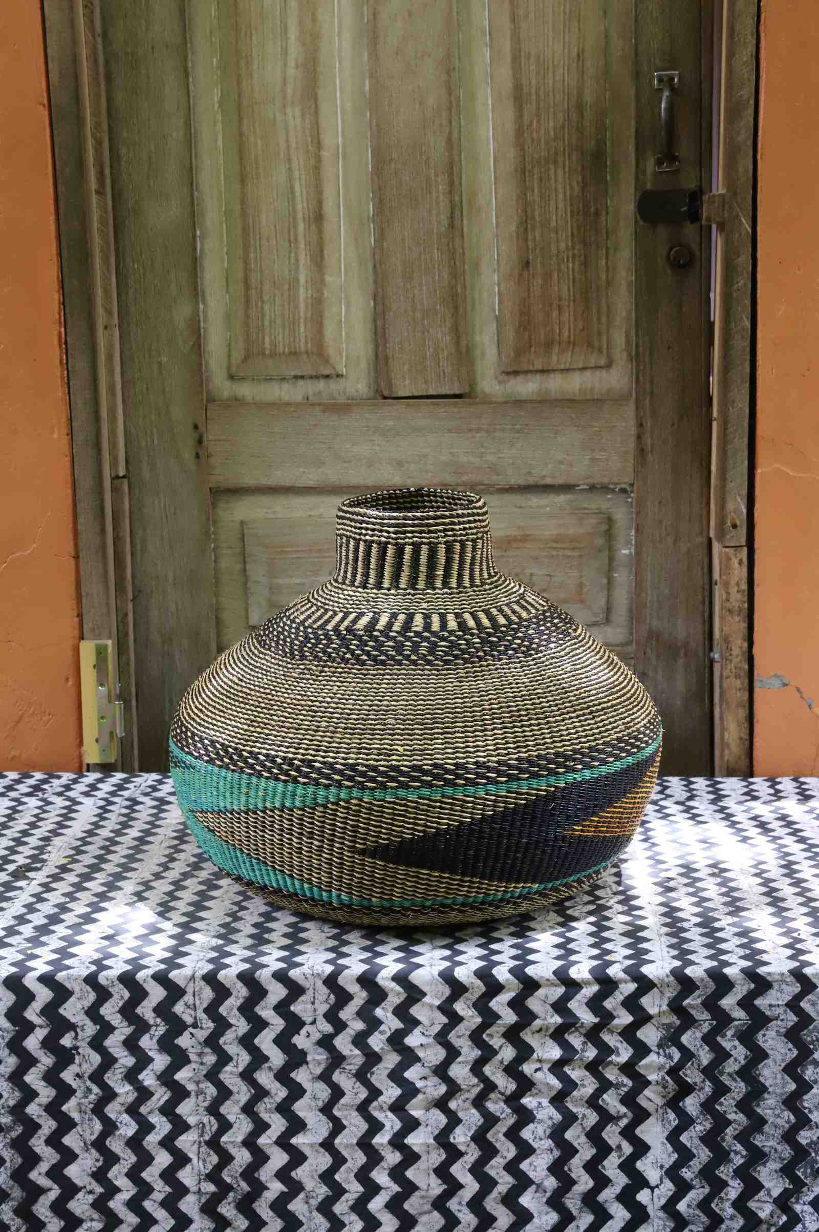 Handwoven African art basket in vase form, natural straw with black and teal geometric pattern, on a chevron surface.