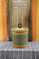Handwoven Bolga laundry basket with coiled rim and lid in natural straw, striped in yellow-orange and teal.
