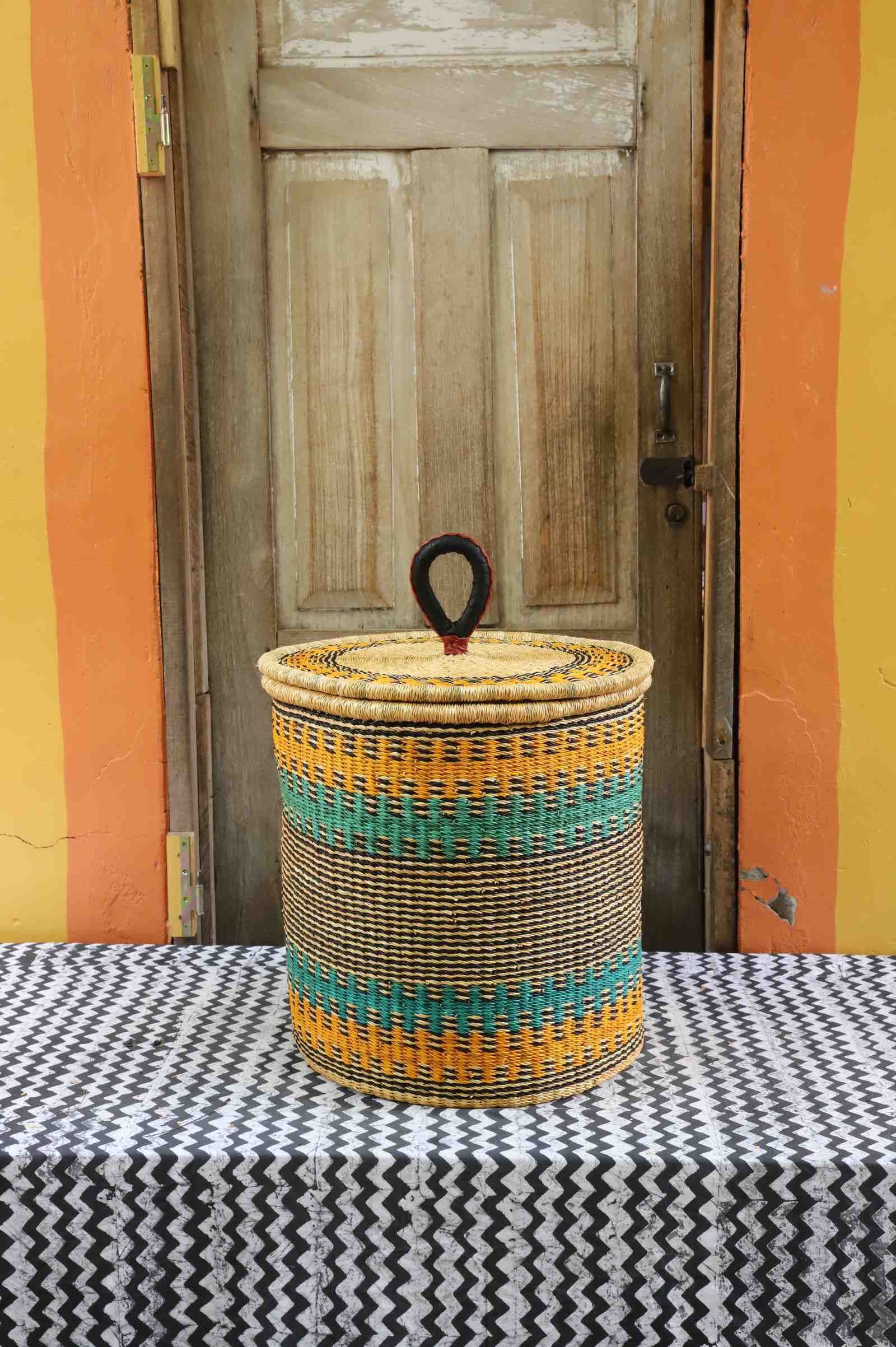 Handwoven Bolga laundry basket with coiled rim and lid in natural straw, striped in yellow-orange and teal.