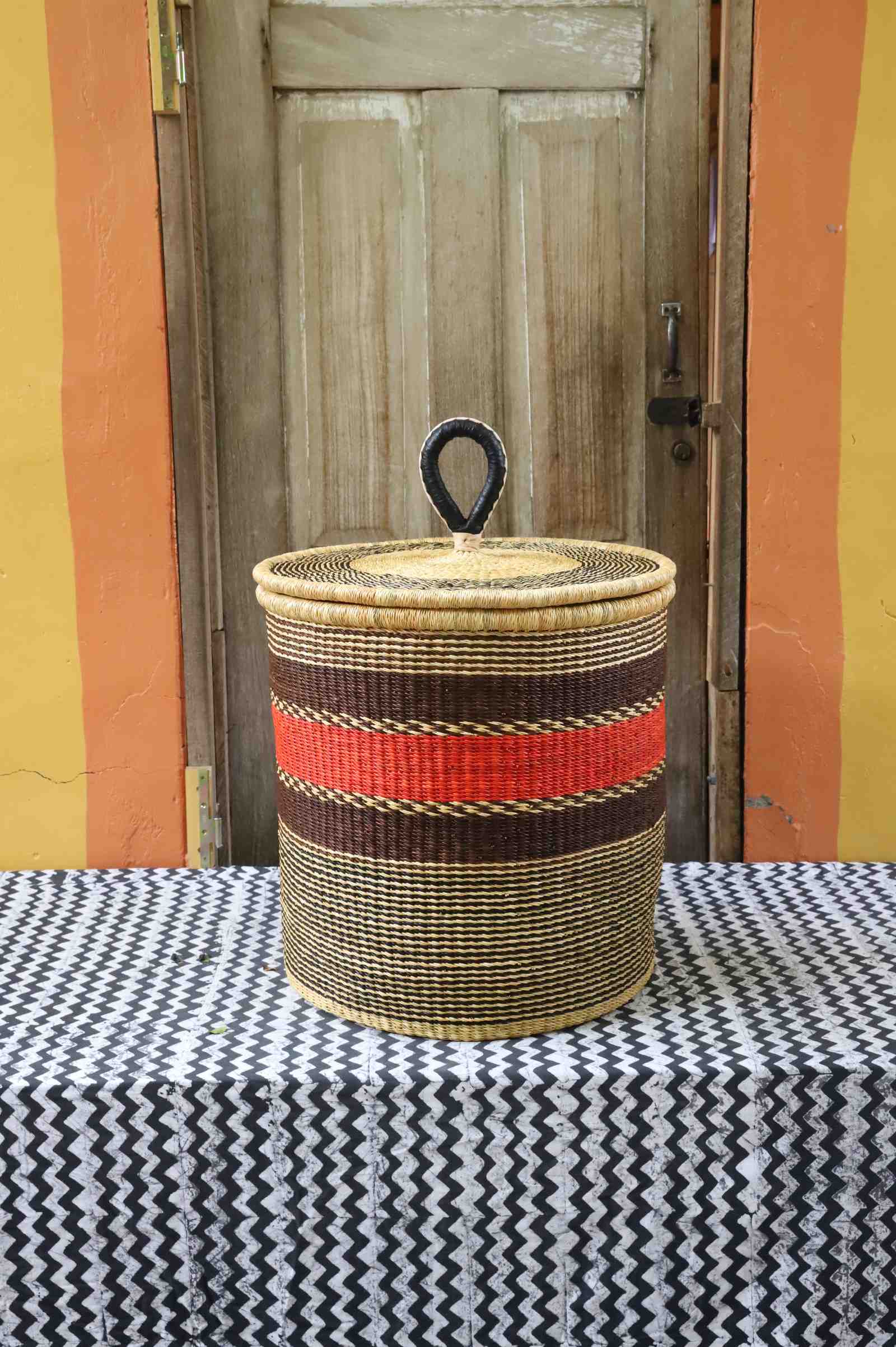 Large coiled-rim lidded laundry basket in natural straw with a red and brown band, styled on a chevron cloth.