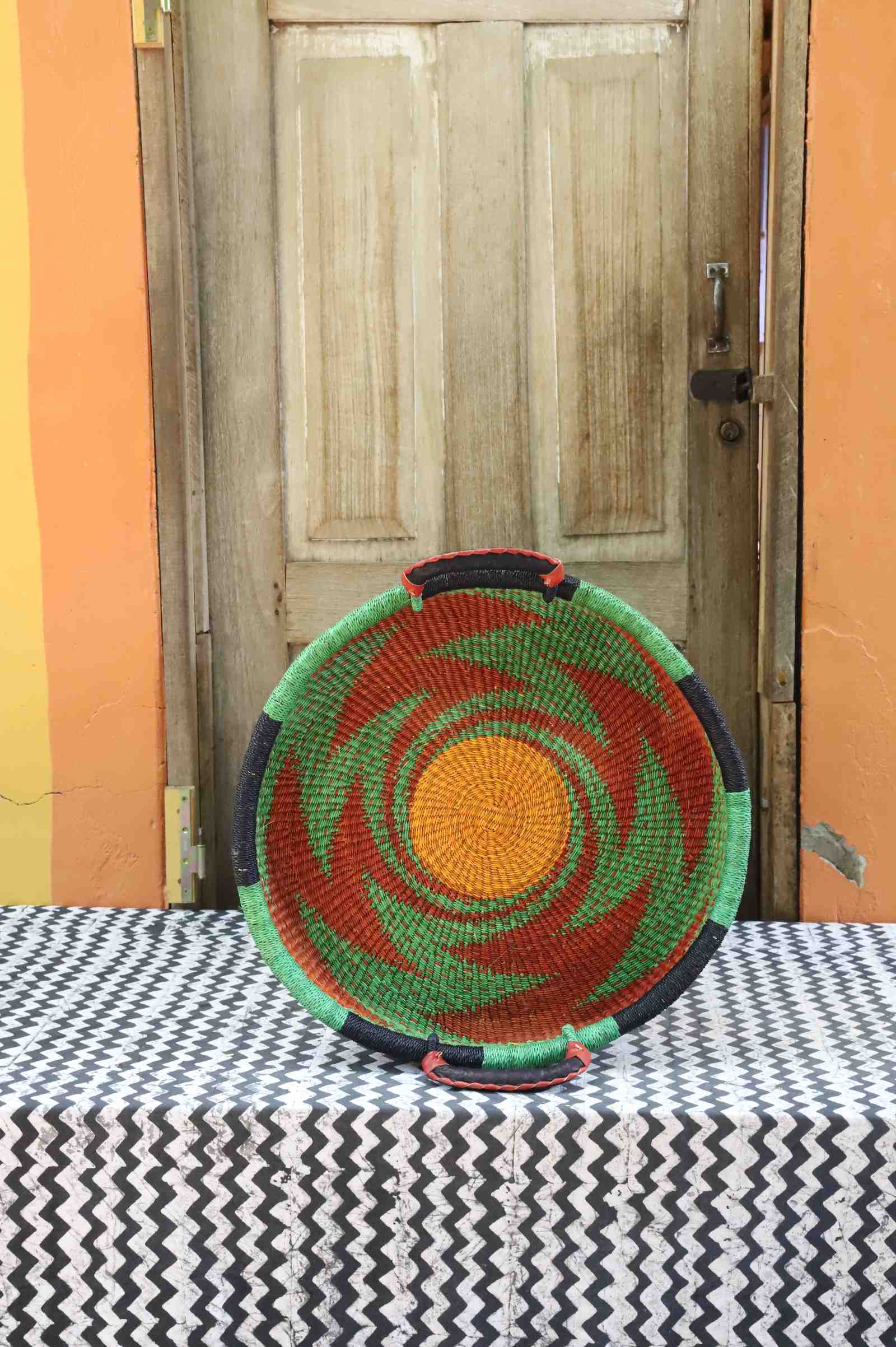 Handwoven Bolga wall tray disc with a green rim and orange pinwheel swirl around a yellow centre.