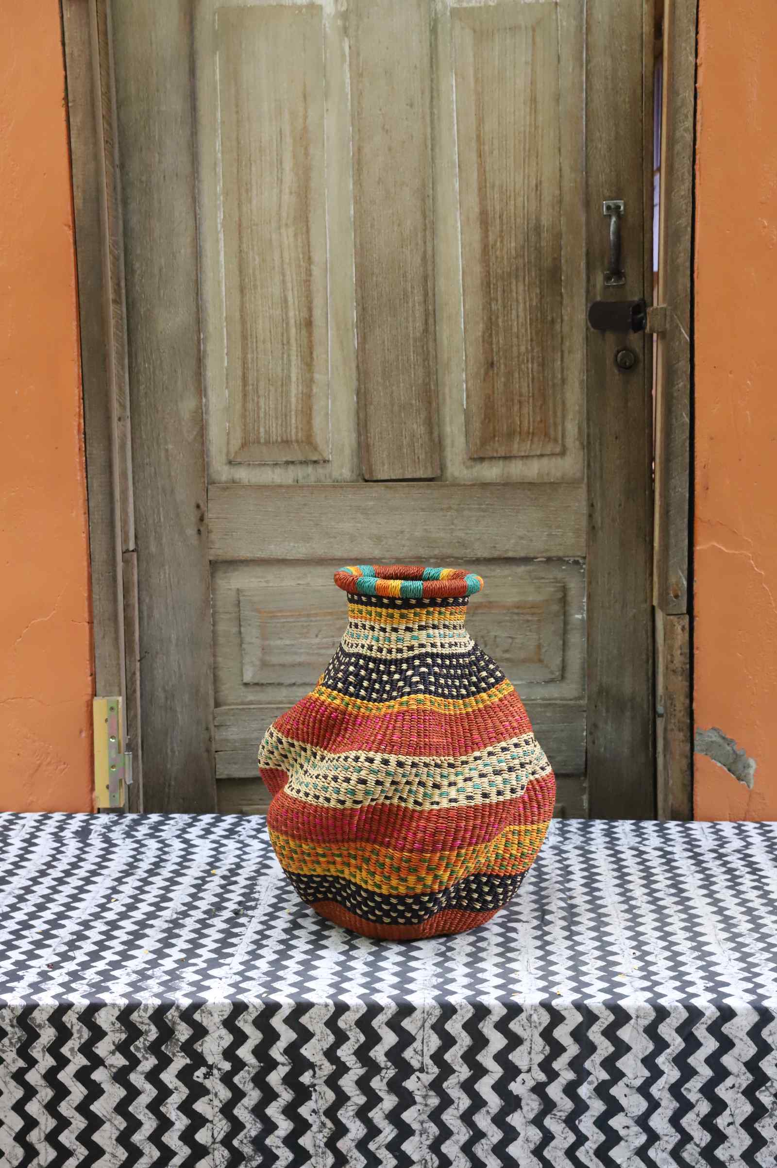 Handwoven Bolga art basket in an urn shape, rust-orange base with black-and-white and orange banded shoulder.