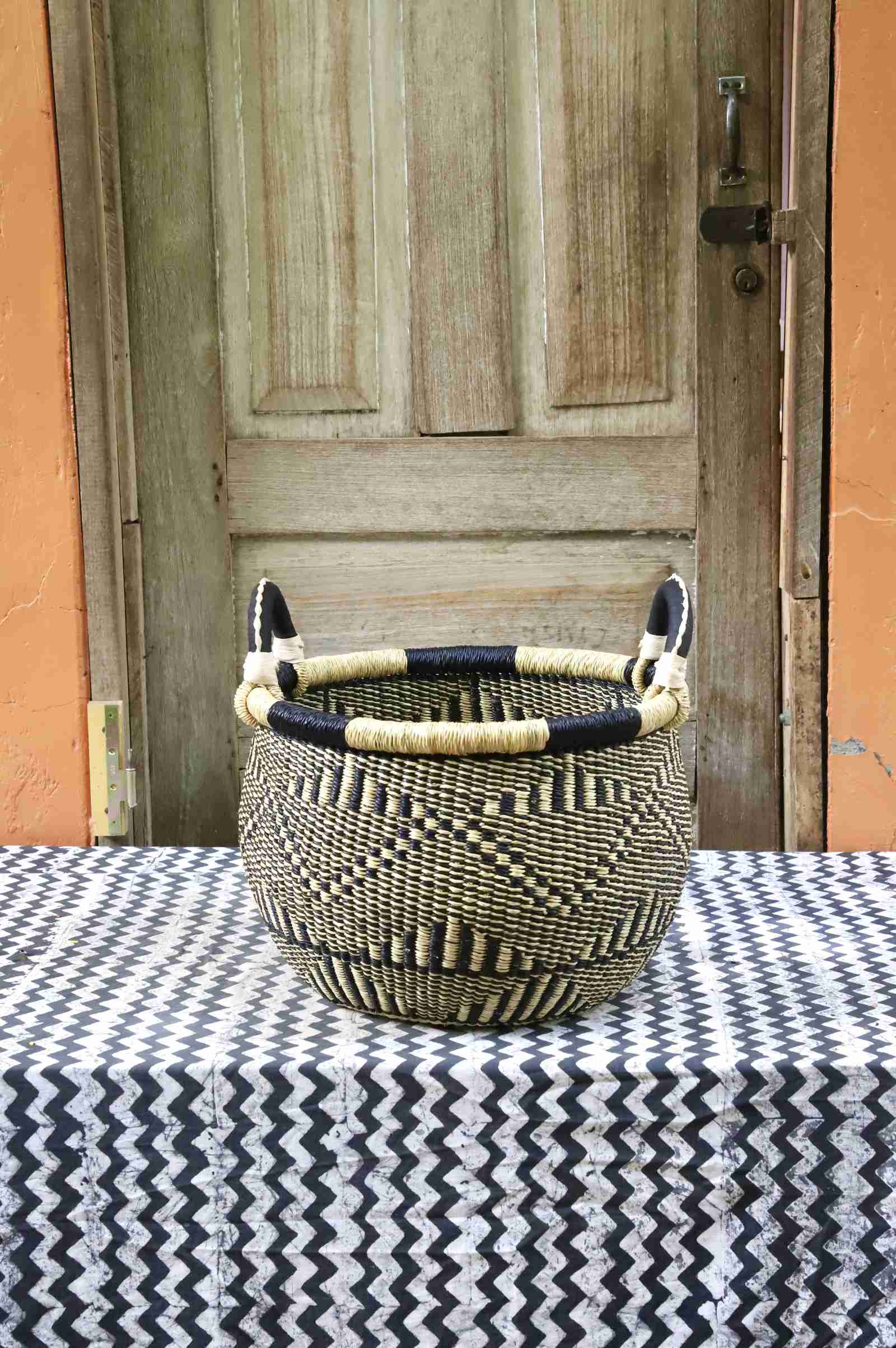 Handwoven Bolga storage basket in natural straw with black diamond pattern and leather loop handles, on a chevron surface.