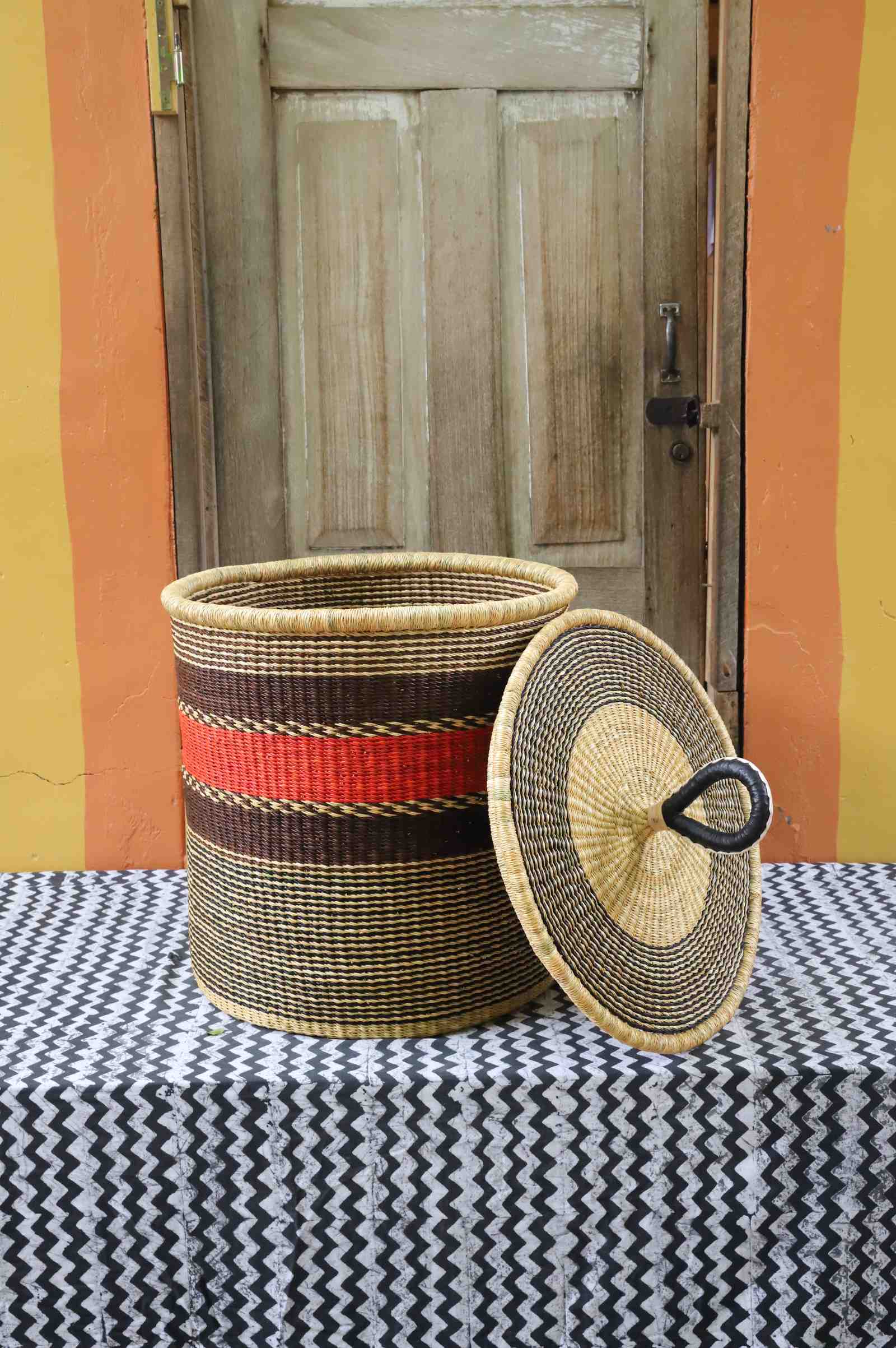 Large coiled-rim laundry basket in natural straw with a red and brown band, lid resting alongside on a chevron cloth.