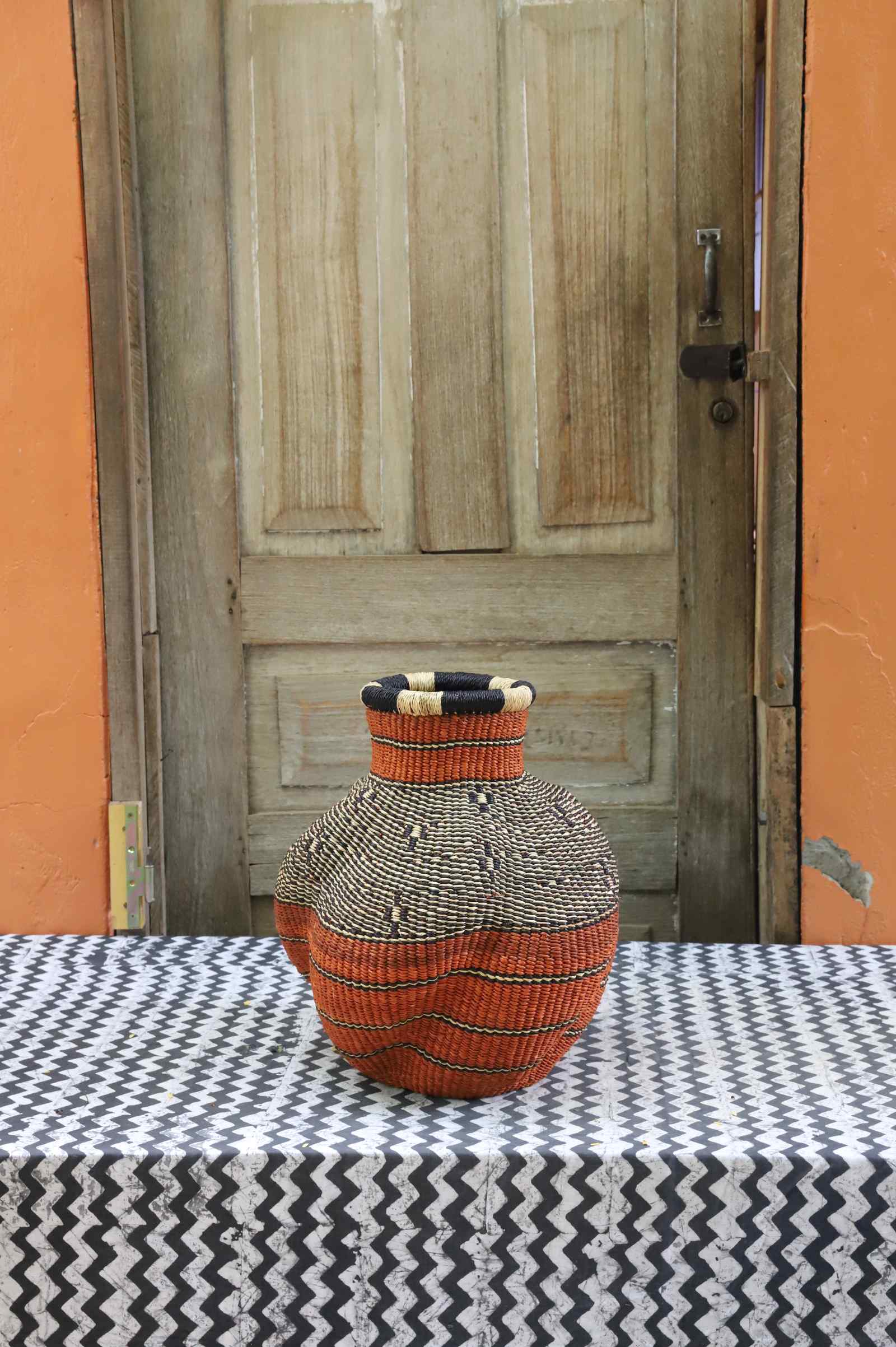 Handwoven Bolga art basket in an urn shape, rust-orange body with a spotted dark shoulder and multicolour rim.