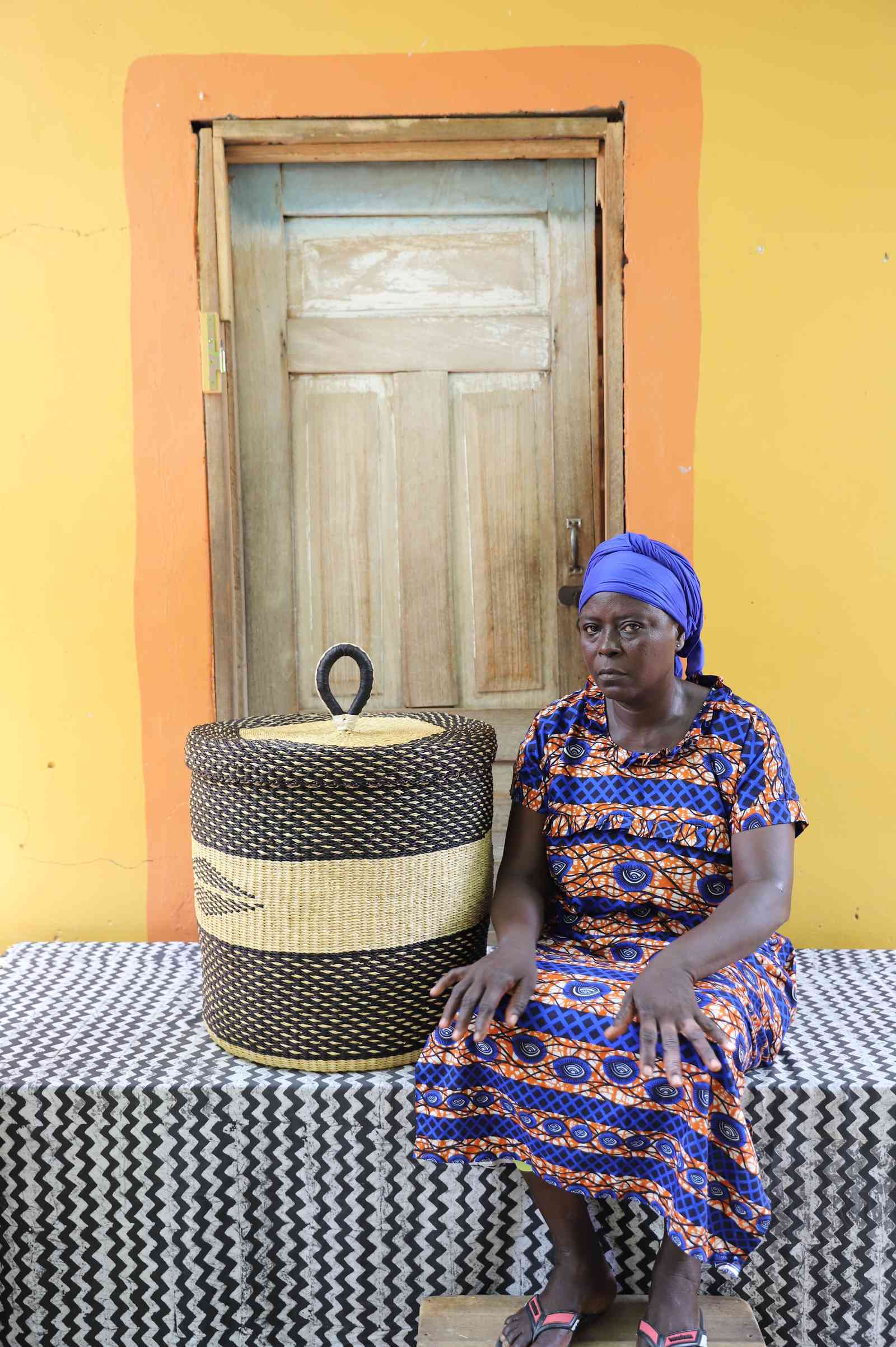 Handwoven Bolga laundry basket in dark straw with natural band, beside a seated woman in a printed dress.