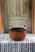 Handwoven Bolga storage basket in rust with black rim and leather loop handles, styled on a chevron surface.