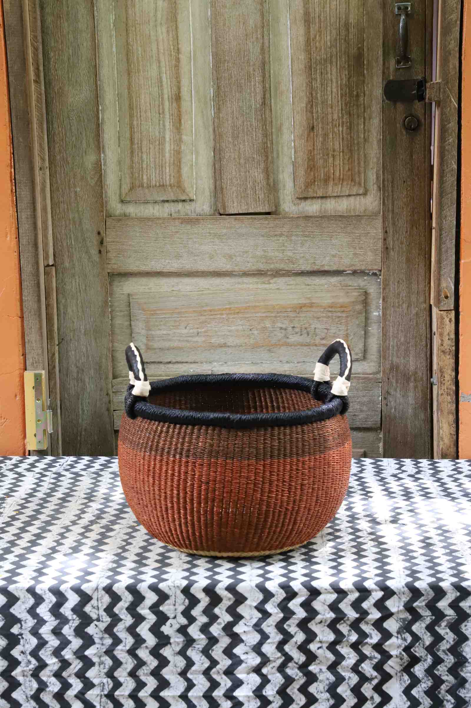 Handwoven Bolga storage basket in rust with black rim and leather loop handles, styled on a chevron surface.
