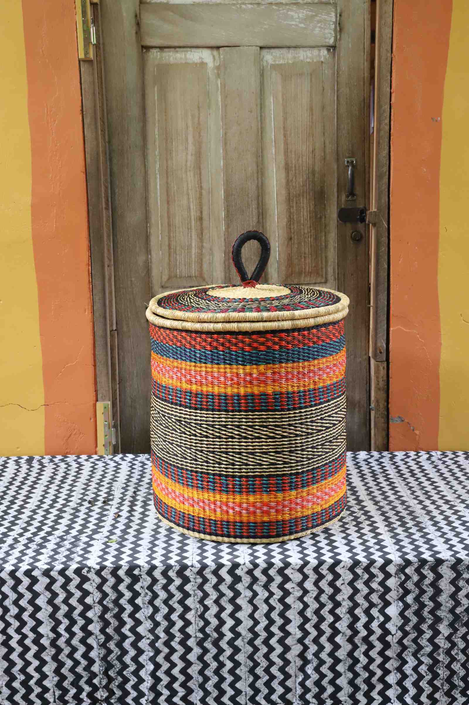 Large coiled-rim lidded laundry basket in rust, teal and gold striped weave, styled on a chevron cloth.