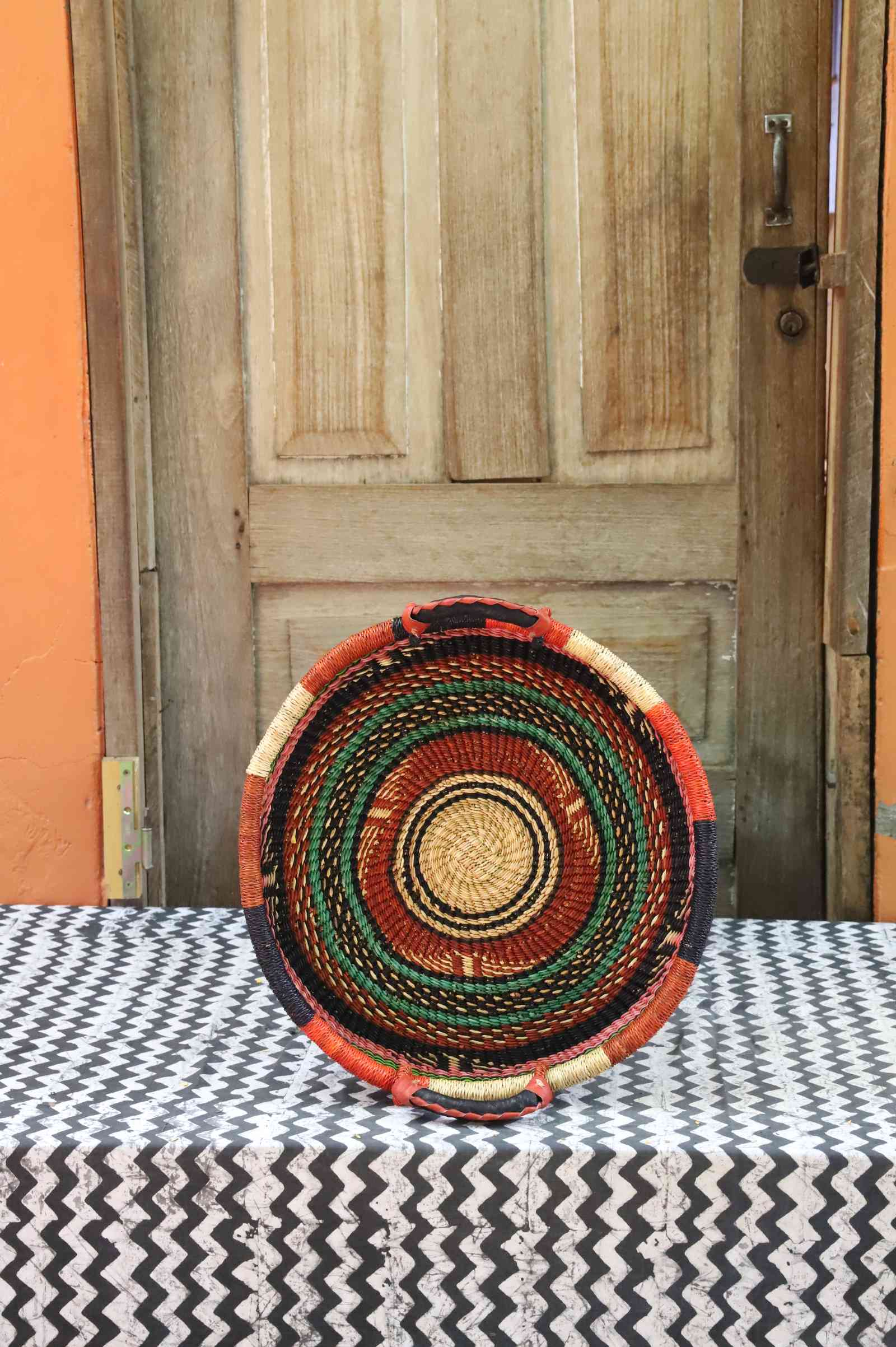 Handwoven Bolga wall tray disc with concentric orange, black, red and green banded rings.