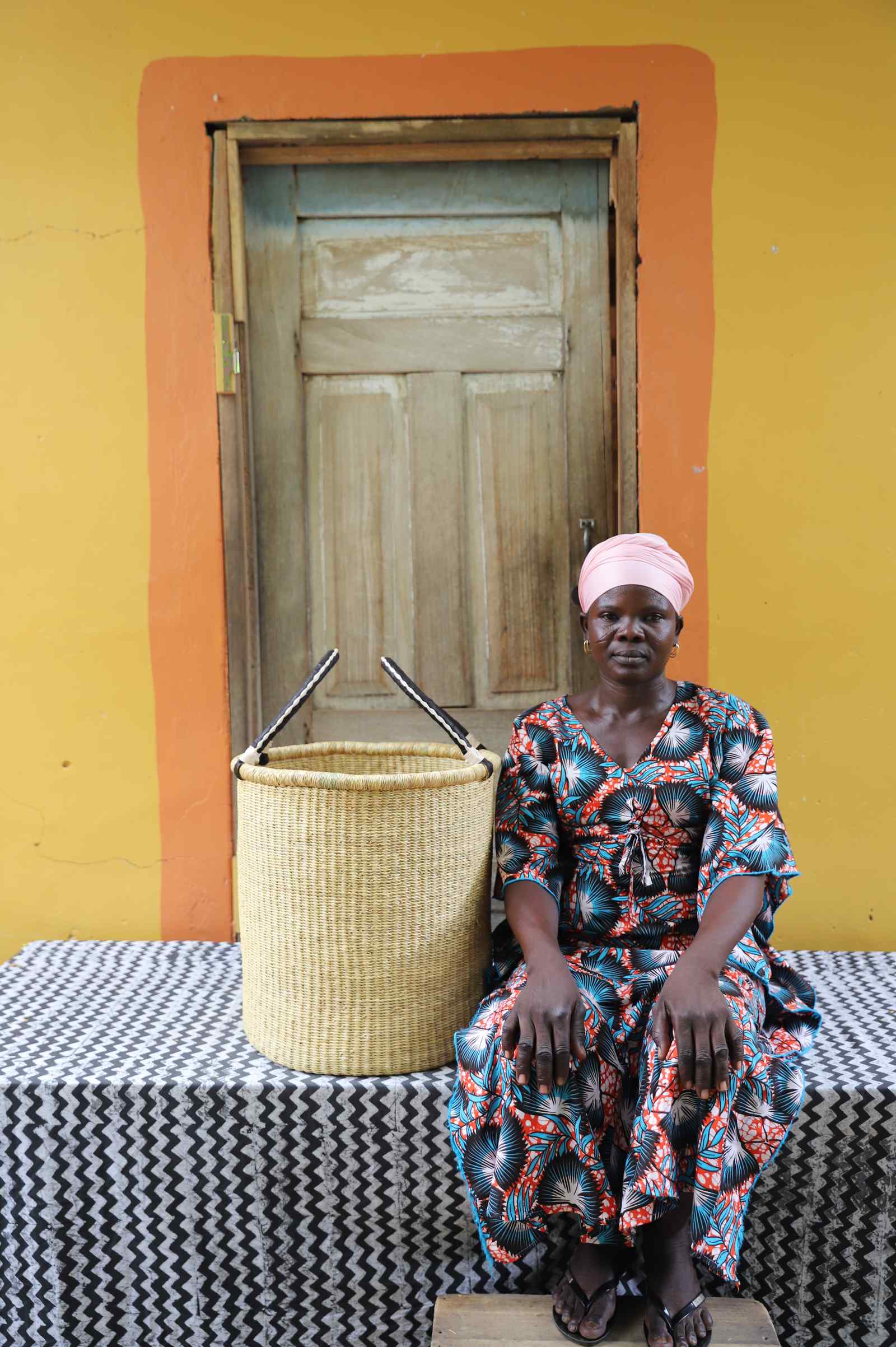 Small handwoven Bolga laundry basket in plain natural straw, beside a seated woman in a printed dress.