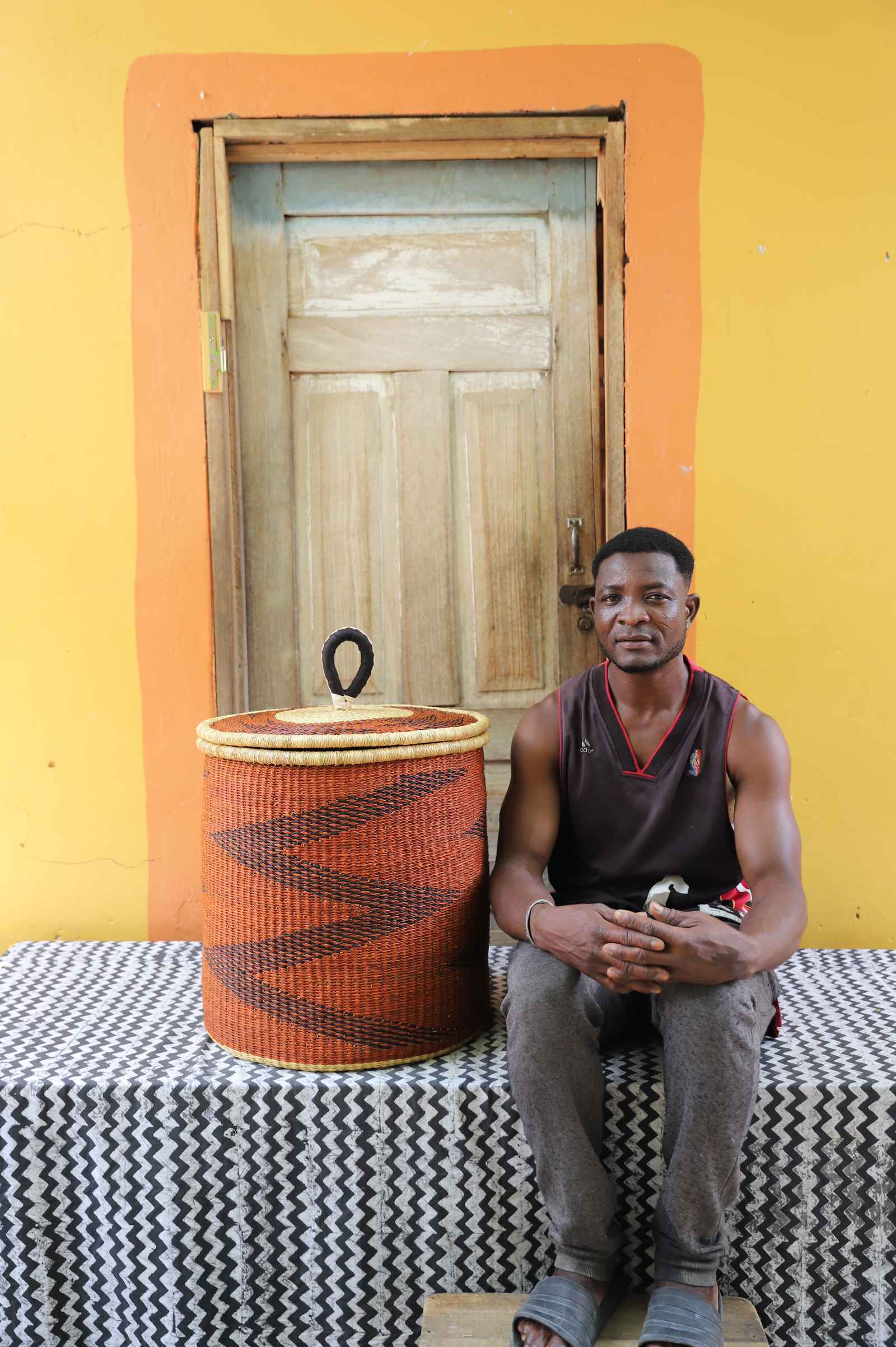 Handwoven Bolga laundry basket in dyed rust-orange with dark zigzag, beside a seated man in a dark vest.