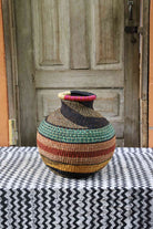Handwoven African décor basket with horizontal bands of gold, terracotta, teal and black, on a chevron surface.