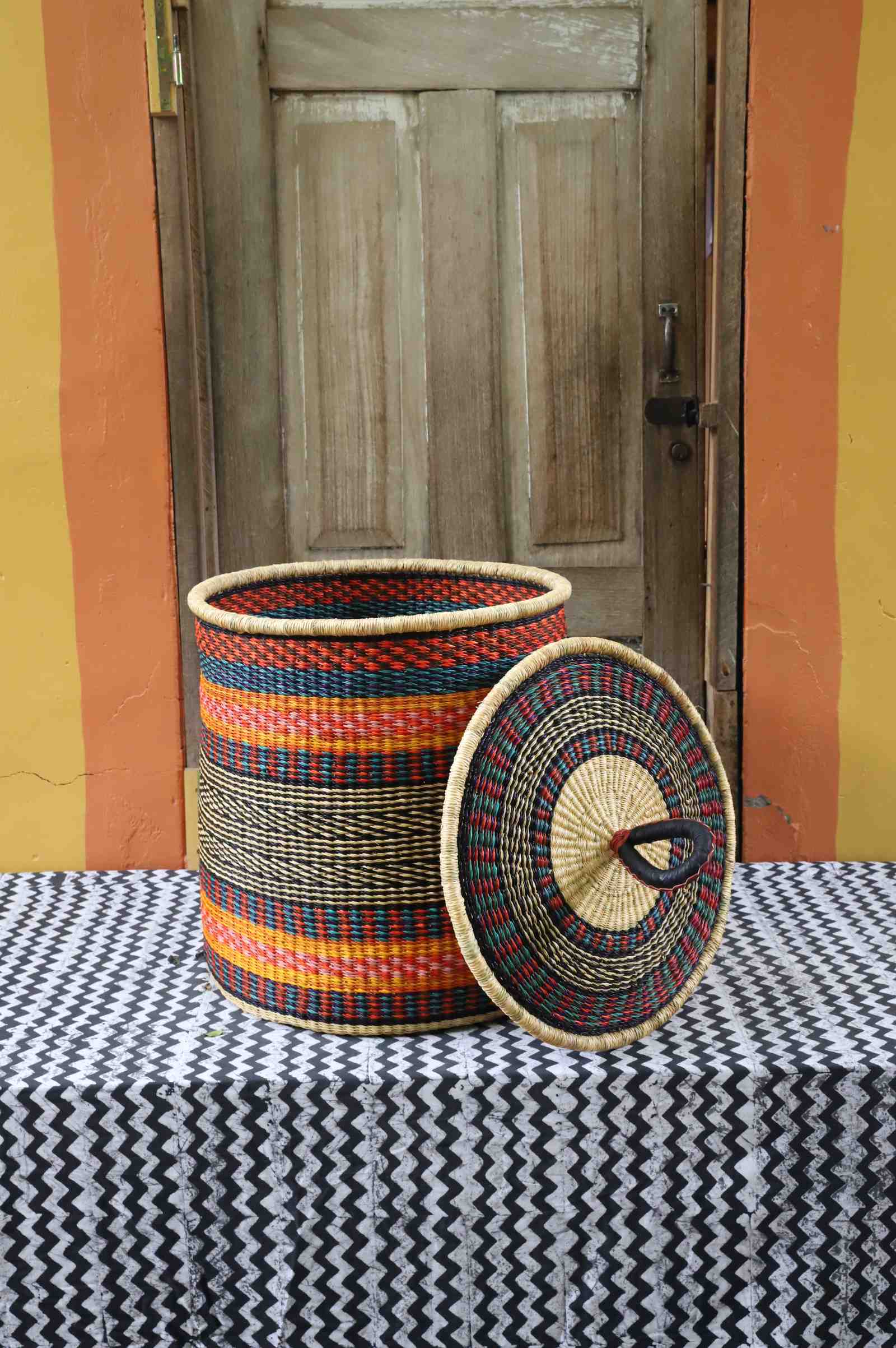 Large coiled-rim laundry basket in rust, teal and gold striped weave, lid resting alongside on a chevron cloth.