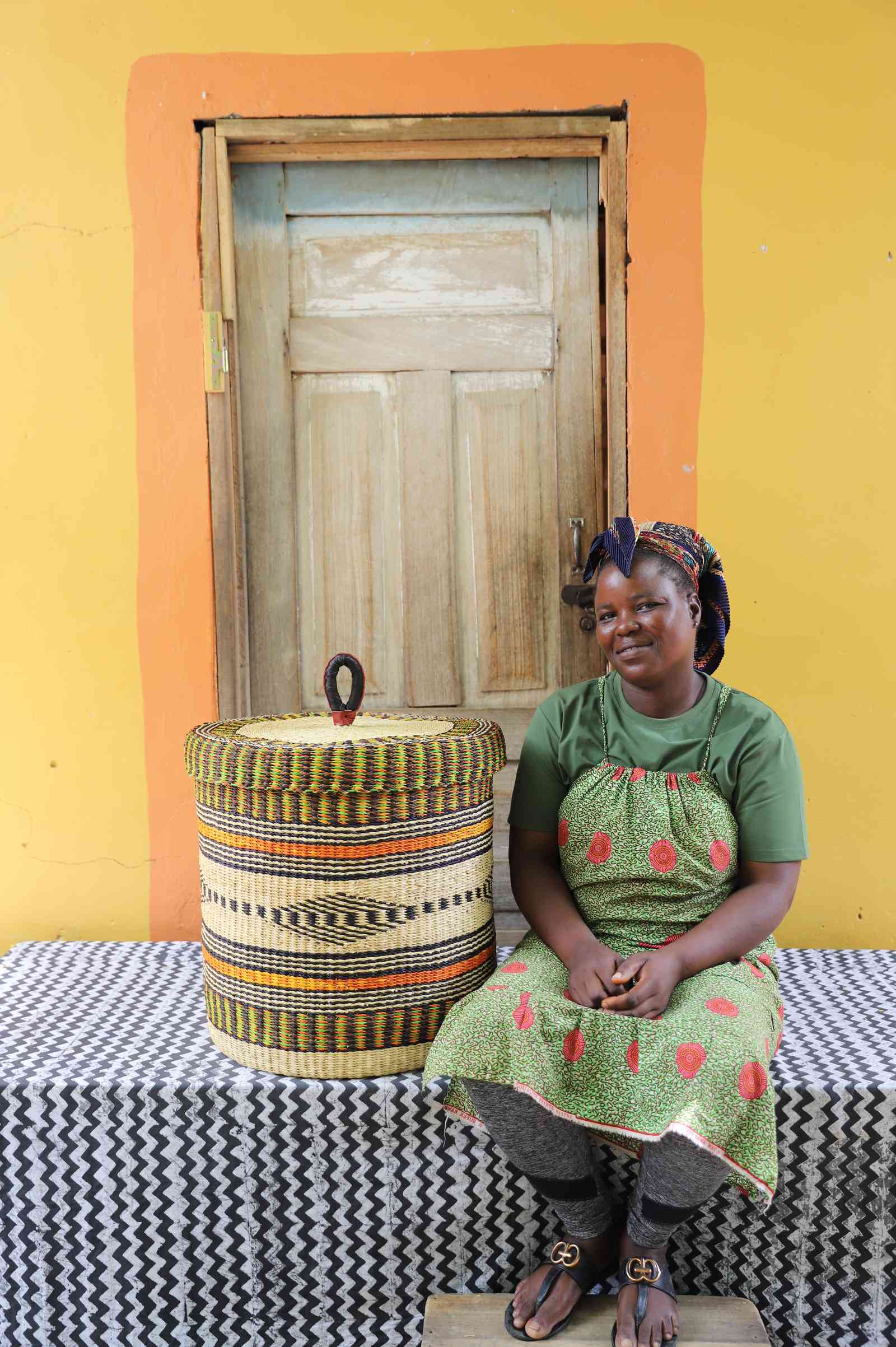 Handwoven Bolga laundry basket in multicolour bands, styled beside a seated woman in a printed dress.