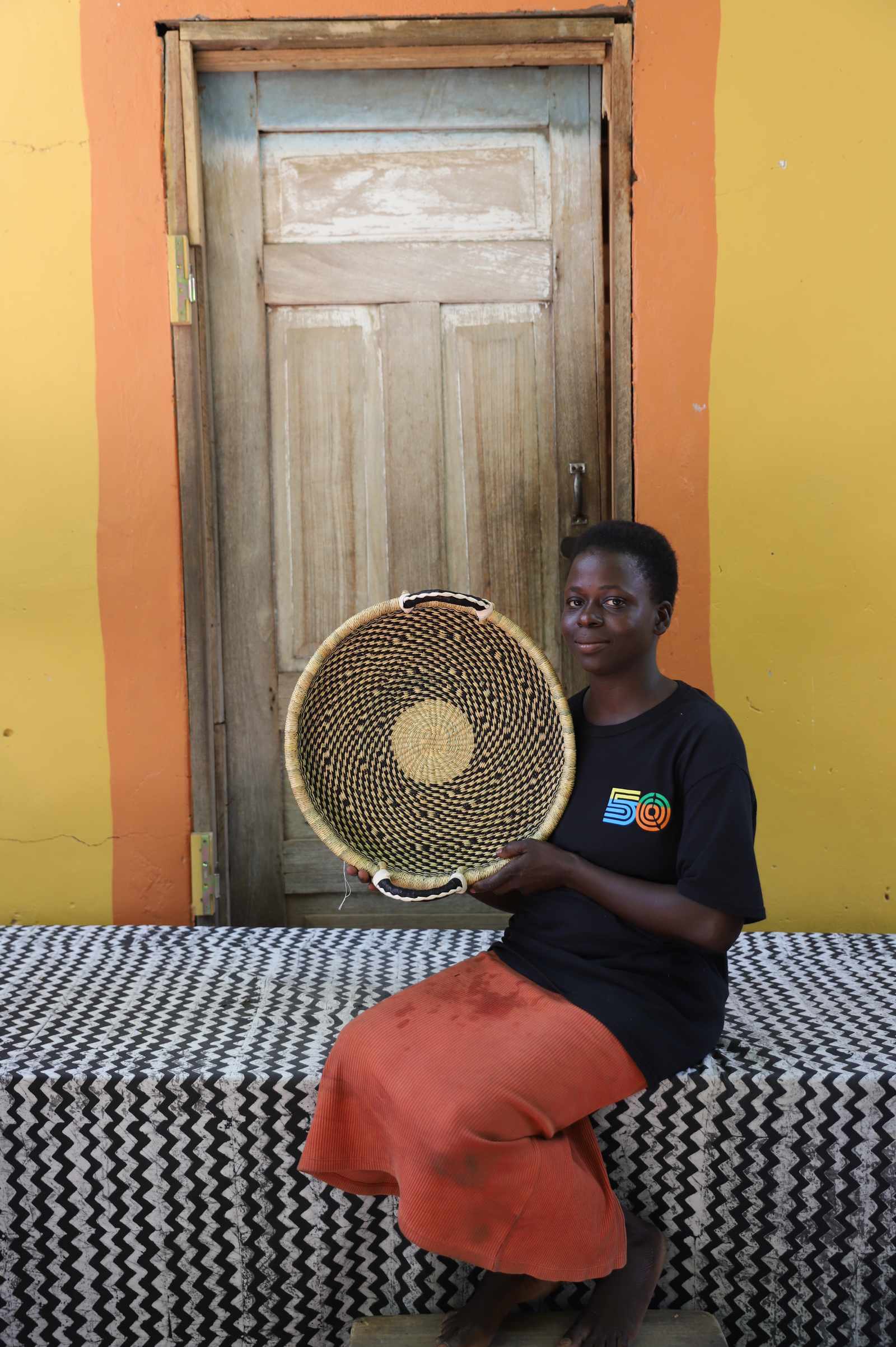 Handwoven Bolga wall tray disc in dark straw with yellow centre, held by a seated woman in dark top.