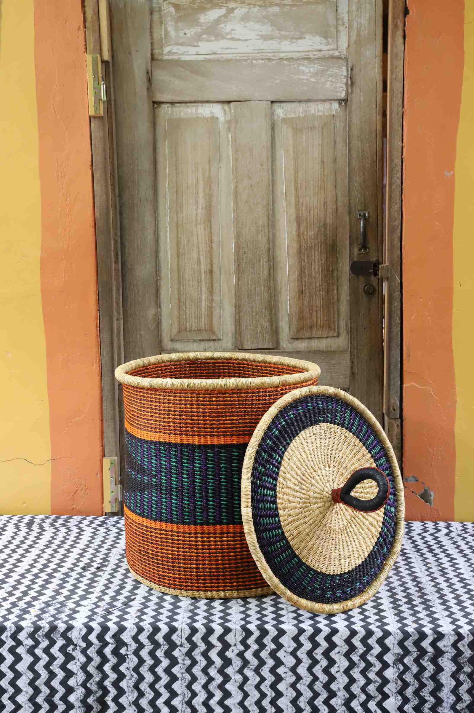 Handwoven Bolga laundry basket with lid removed, natural body with orange and dark blue bands and matching lid.