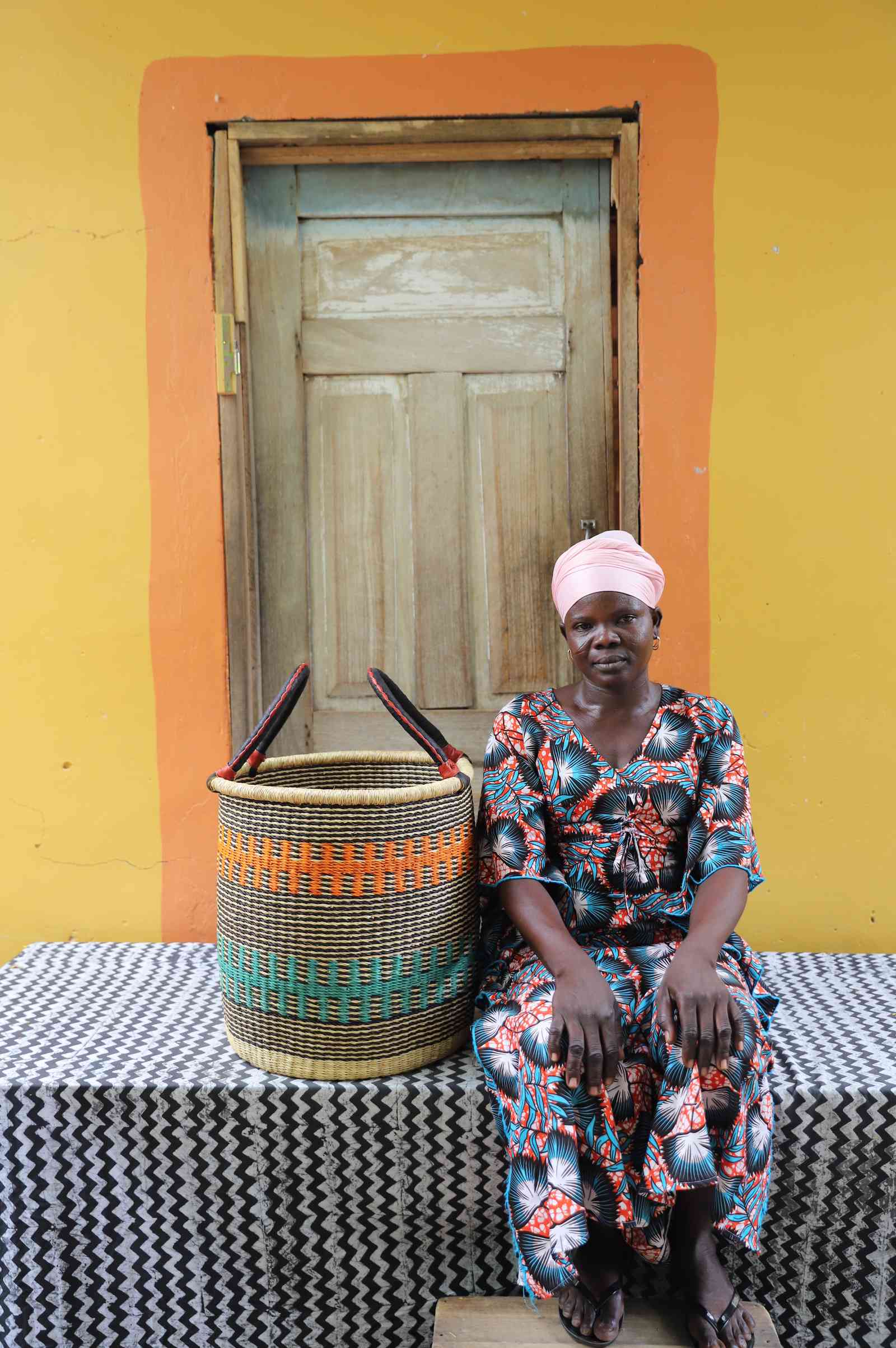 Small handwoven Bolga laundry basket in natural with orange and teal bands, beside a seated woman in floral.