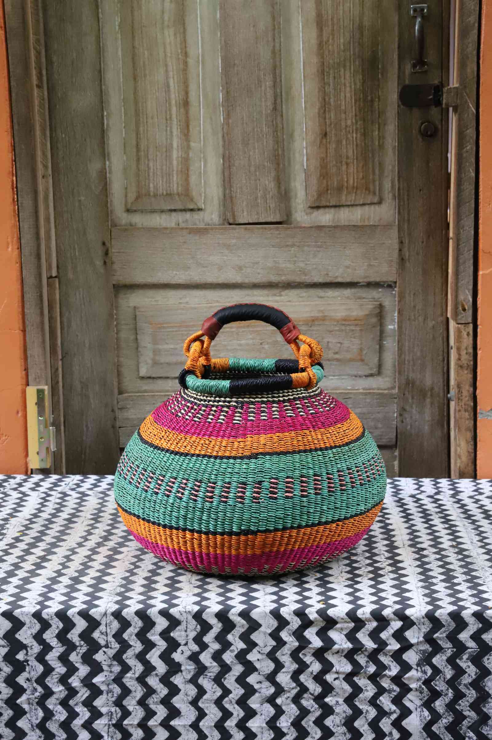 Large handwoven Bolga pot basket in bold bands of pink, teal and amber with a leather top handle, on a chevron surface.