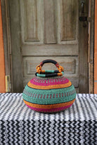 Large handwoven Bolga pot basket in bold bands of pink, teal and amber with a leather top handle, on a chevron surface.