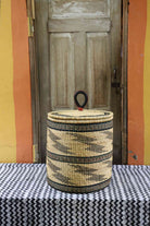Handwoven Bolga laundry basket with coiled rim and lid in natural straw with fine black bands and wave motifs.