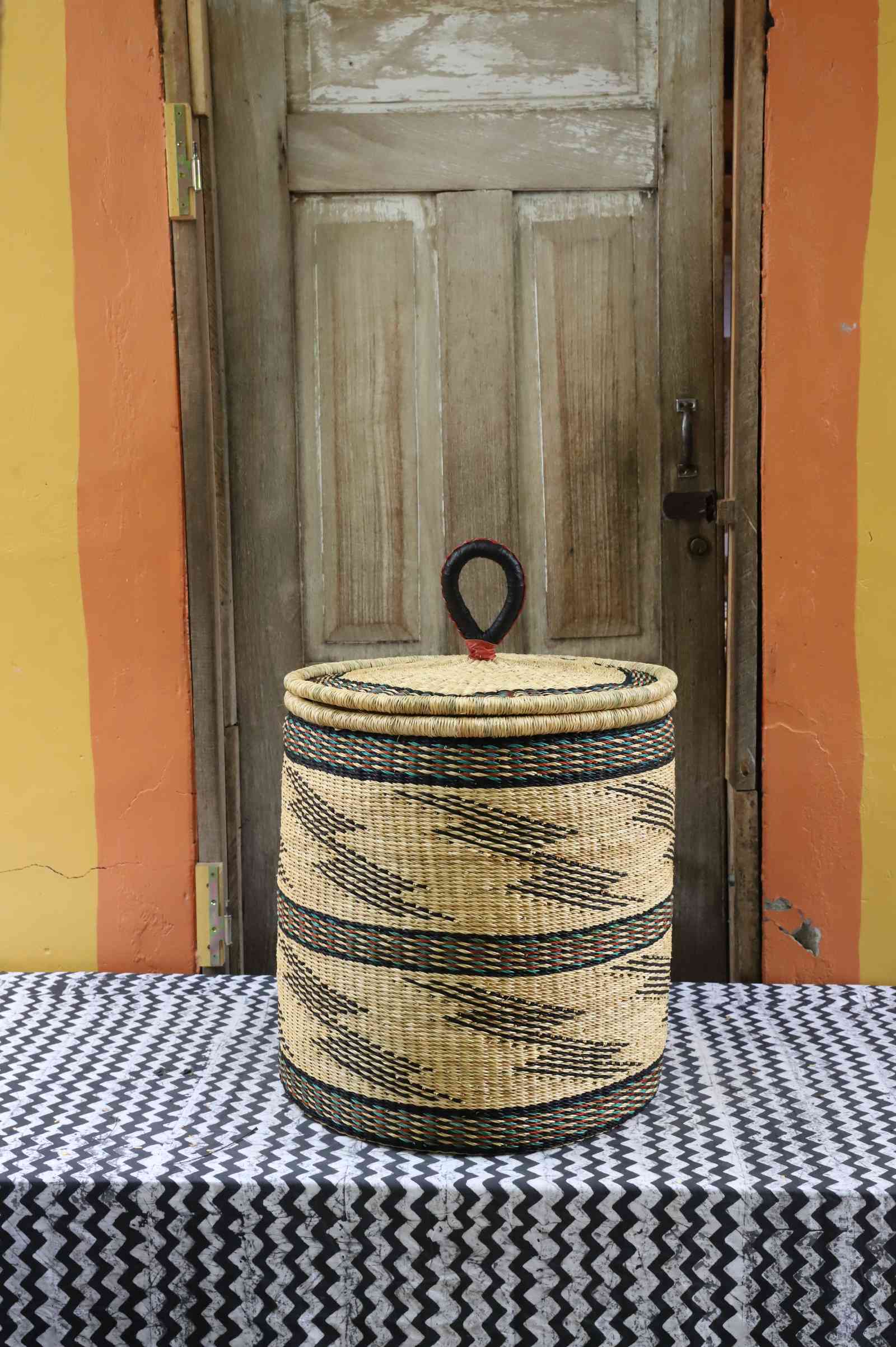 Handwoven Bolga laundry basket with coiled rim and lid in natural straw with fine black bands and wave motifs.