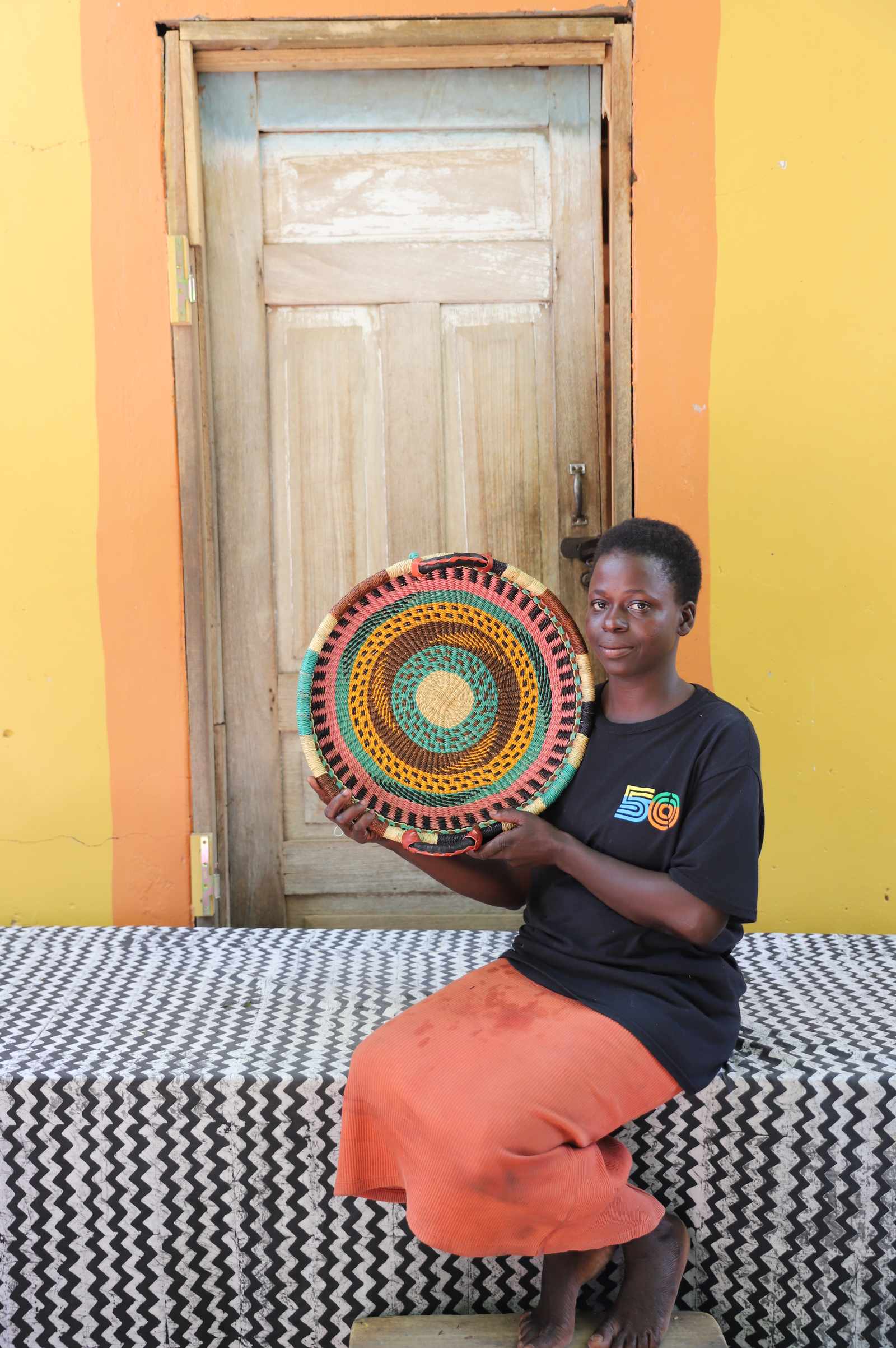 Handwoven Bolga wall tray disc with red and green bands, held by a seated woman in an orange skirt.