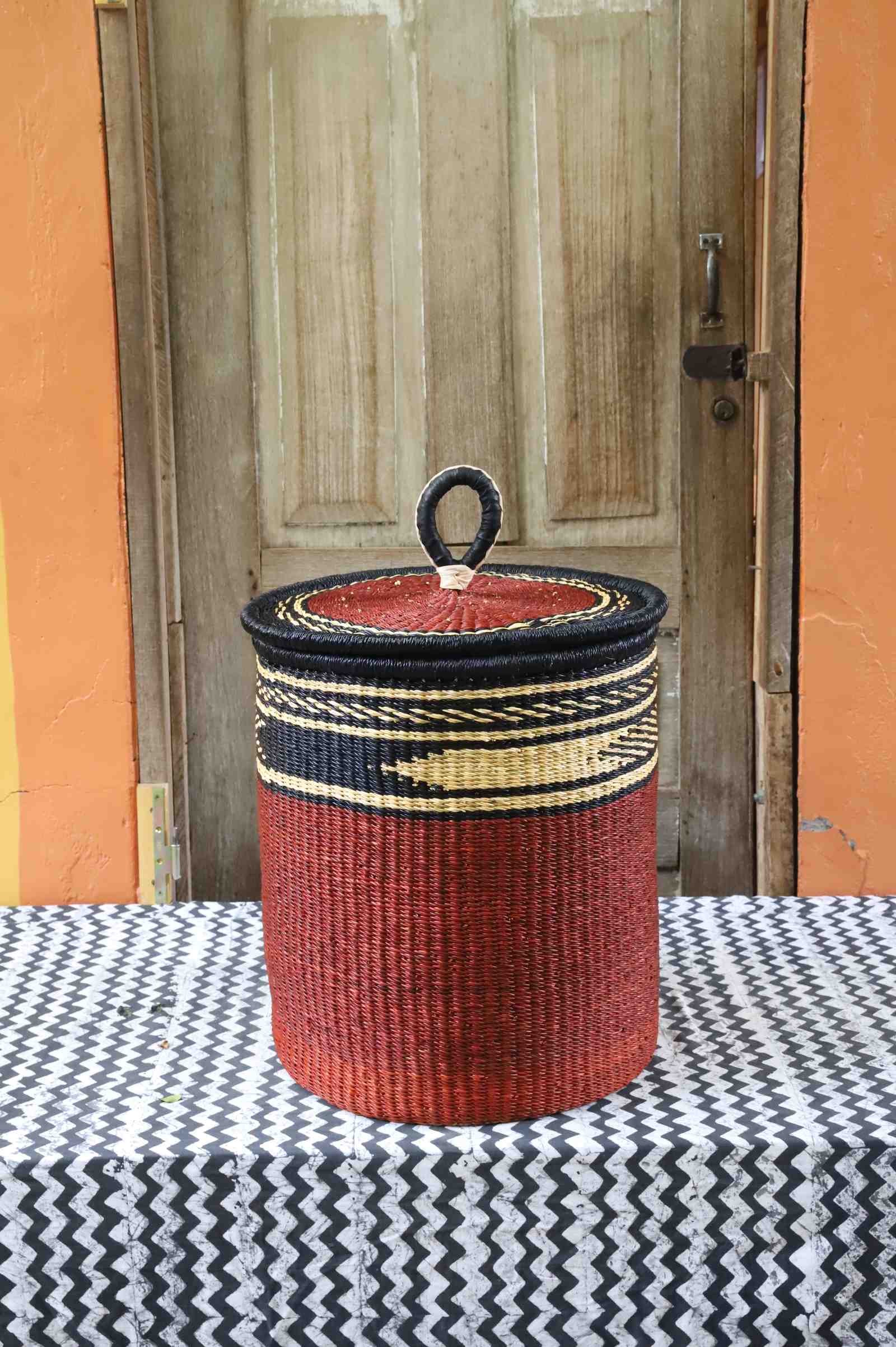 Coiled-rim lidded laundry basket in solid rust weave with a black and tan banded rim, styled on a chevron cloth.