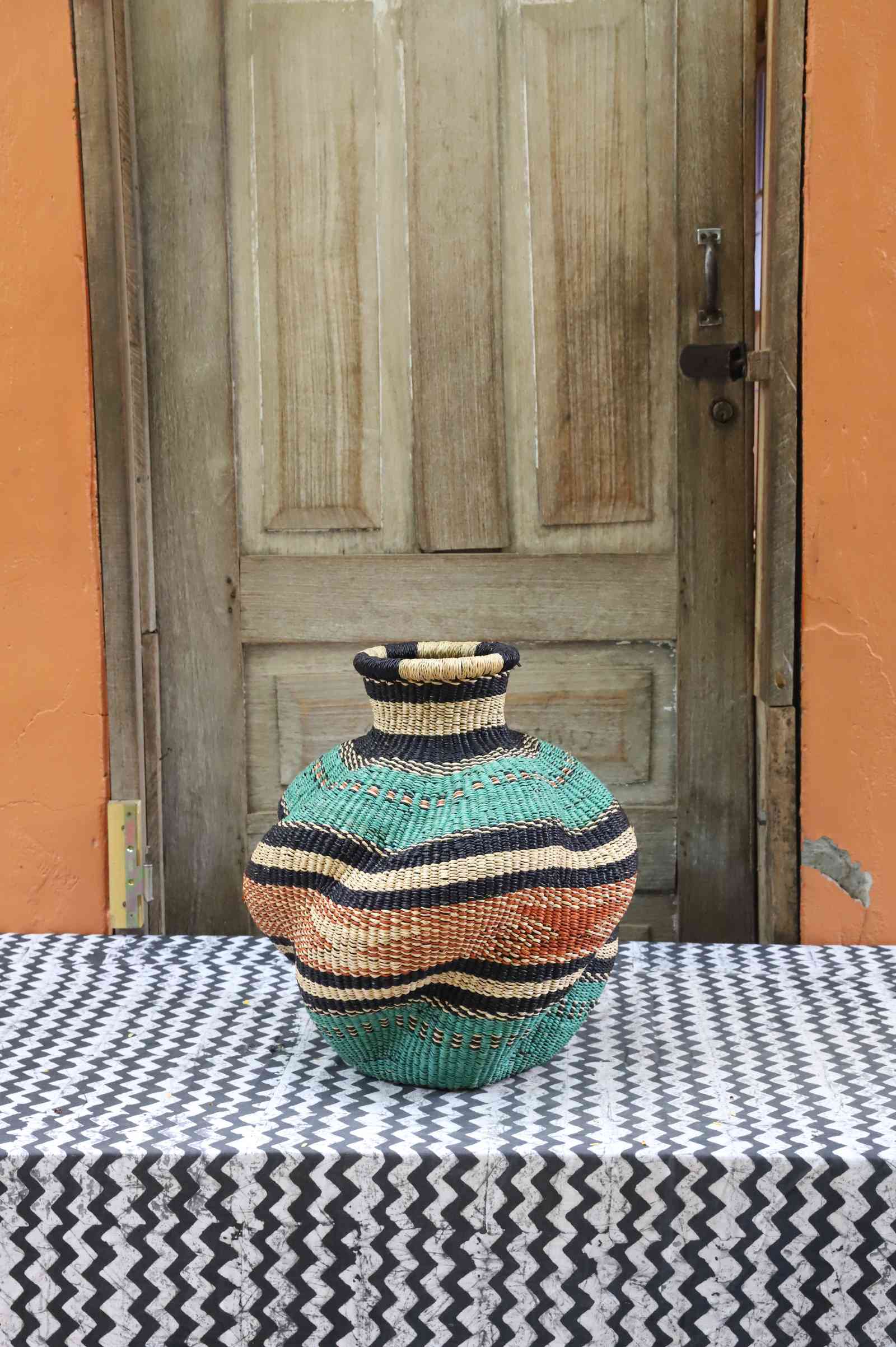 Handwoven Bolga art basket in an urn shape, teal body with pink, orange and black banded shoulder.