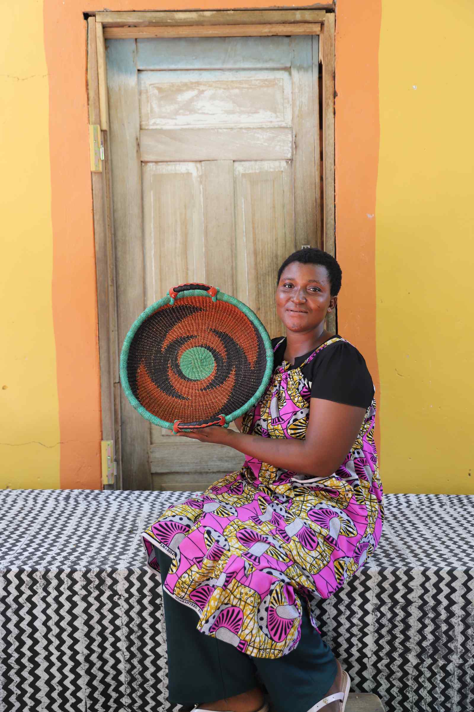 Handwoven Bolga wall tray disc with green rim and orange bands, held by a seated woman in floral.