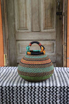 Handwoven Bolga pot basket in multicoloured bands of green, teal and amber with a leather top handle, on a chevron surface.
