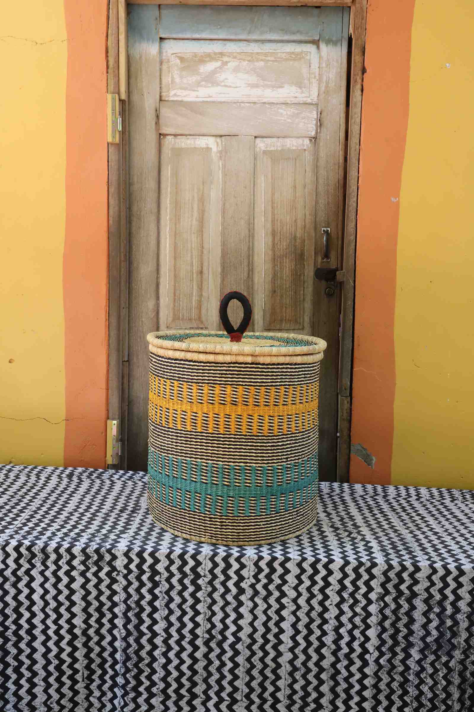 Handwoven Bolga laundry basket with coiled rim and lid in natural straw with yellow and teal banded stripes.