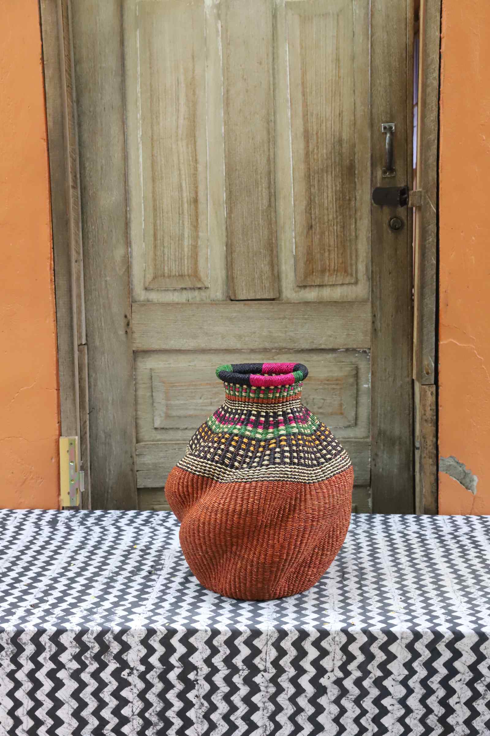 Handwoven Bolga art basket in an urn shape, rust-orange body with dark and multicolour banded shoulder.