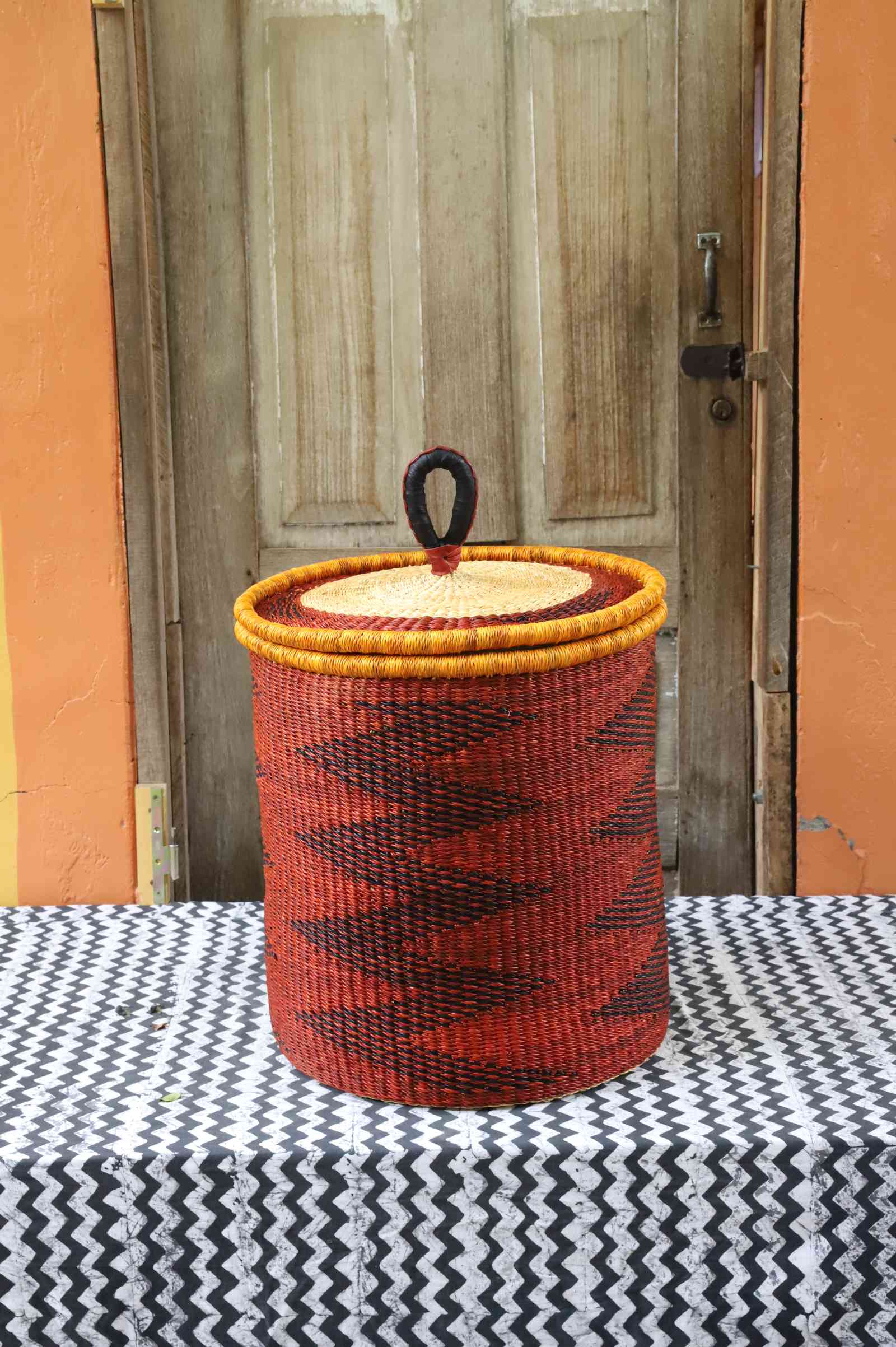 Coiled-rim lidded laundry basket in rust weave with a black diamond pattern and mustard rim, styled on a chevron cloth.