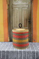 Handwoven Bolga laundry basket with coiled rim and lid in natural straw with red, orange and yellow banded stripes.