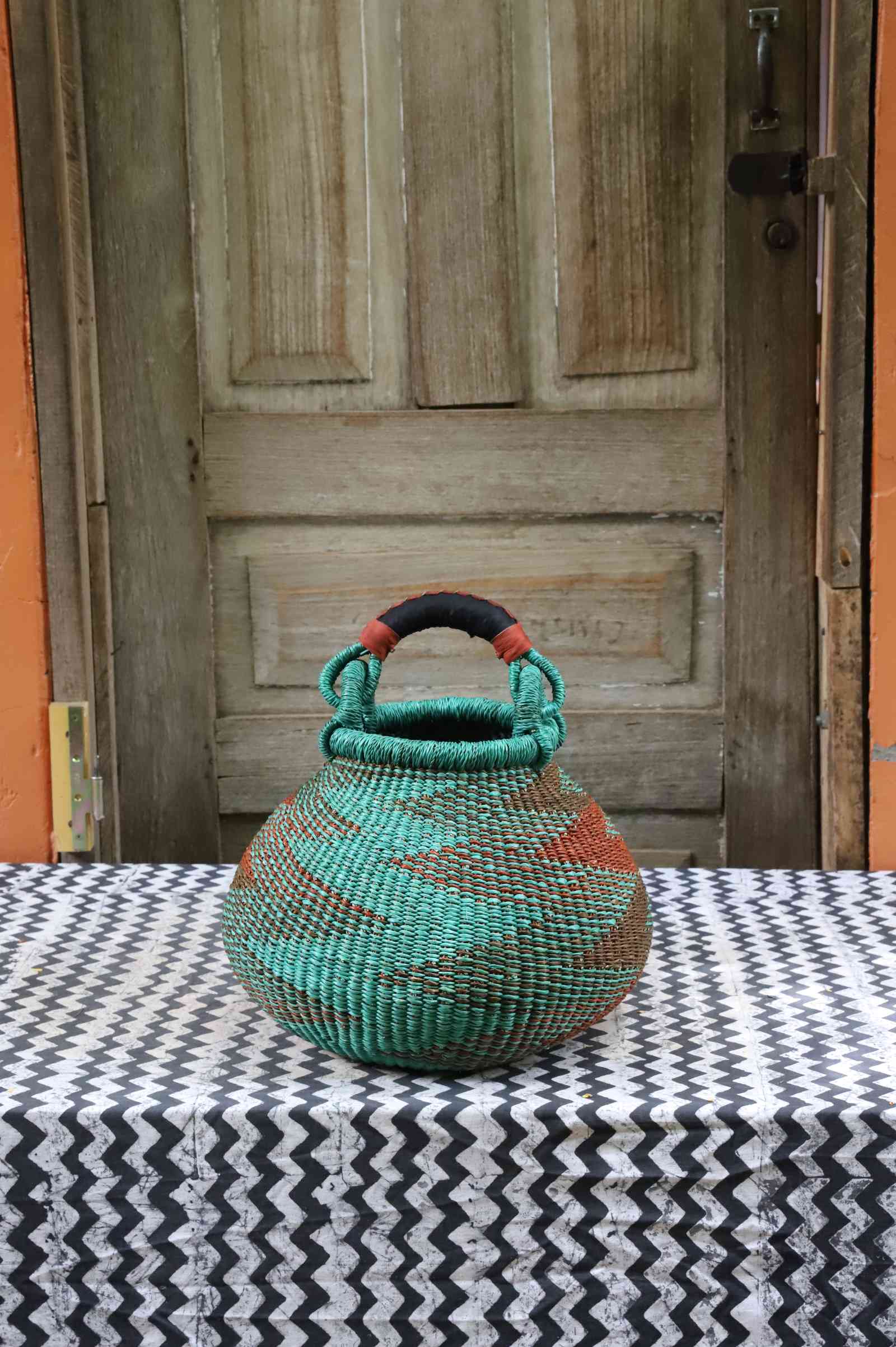 Small handwoven Bolga pot basket in teal and terracotta elephant grass with a leather top handle, on a chevron surface.