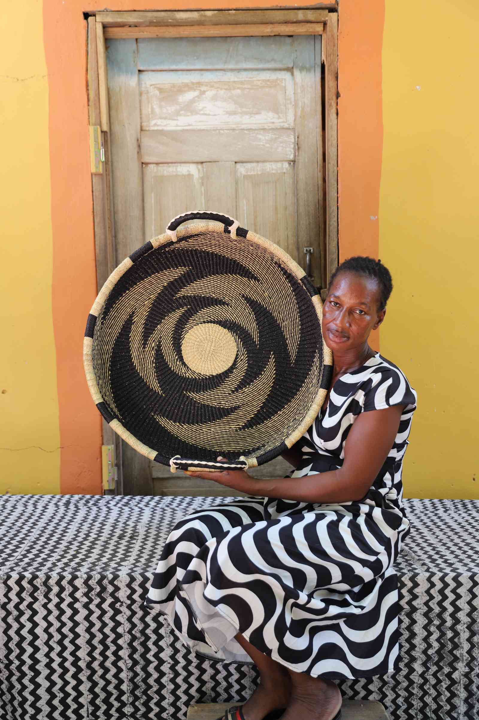 Medium handwoven Bolga wall tray disc with bold black pinwheel, held by a seated woman in stripes.