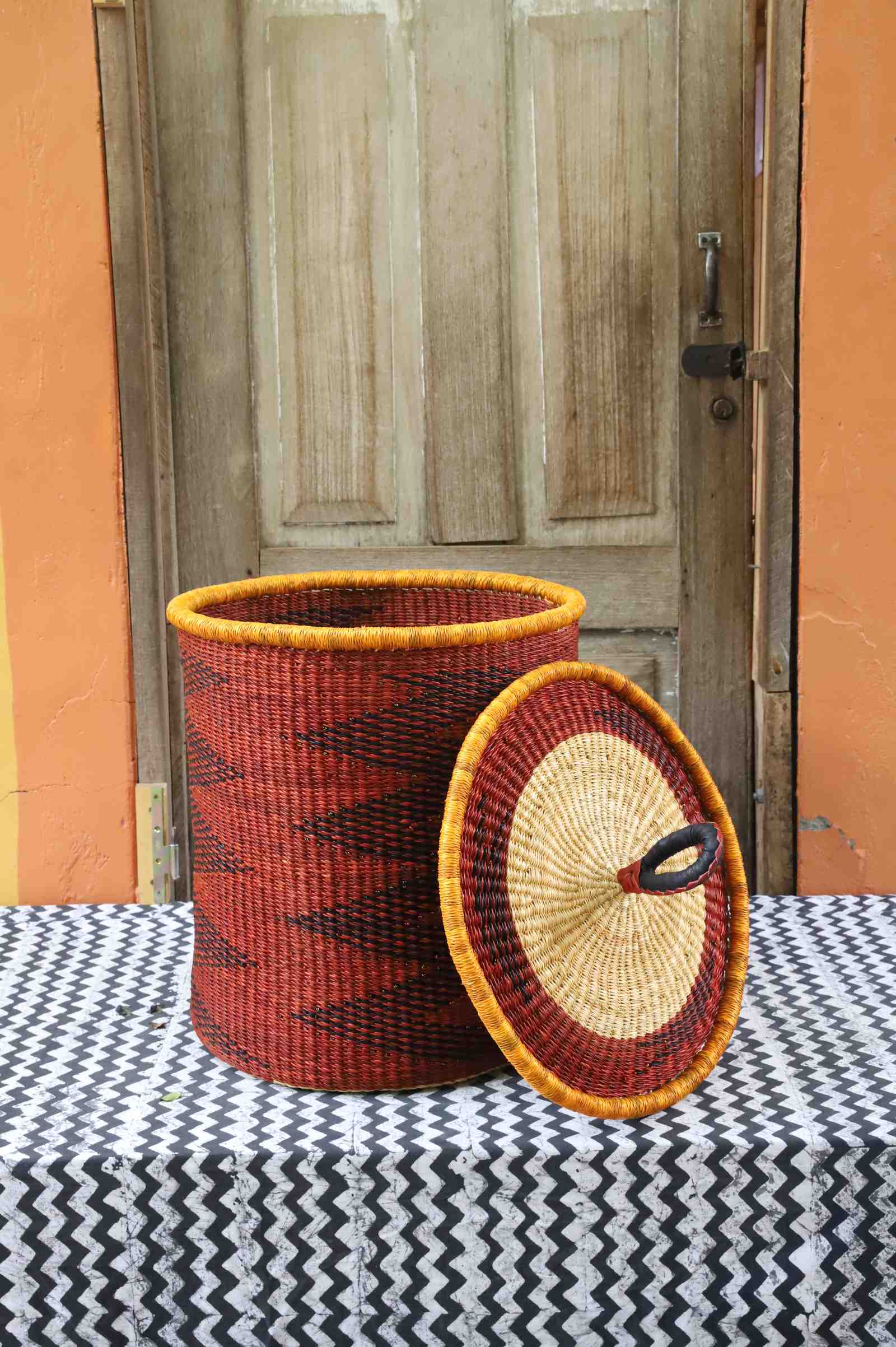 Coiled-rim laundry basket in rust weave with a black diamond pattern, lid resting alongside on a chevron cloth.