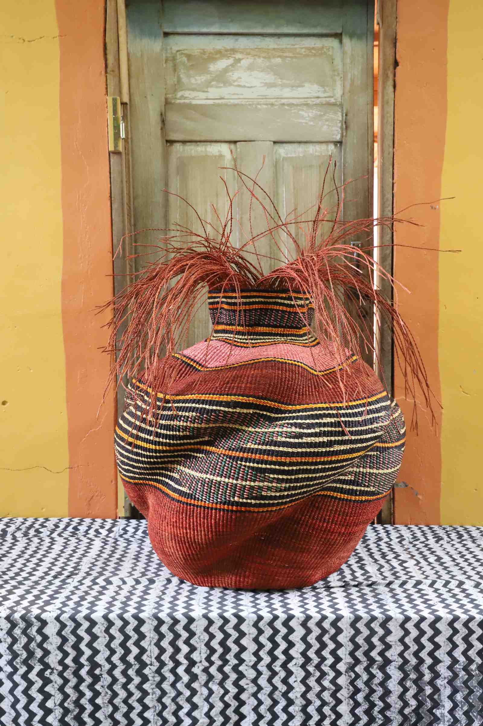 Large round Bolga basket in rust red with a black, cream and orange striped band and loose straw fronds at the neck.