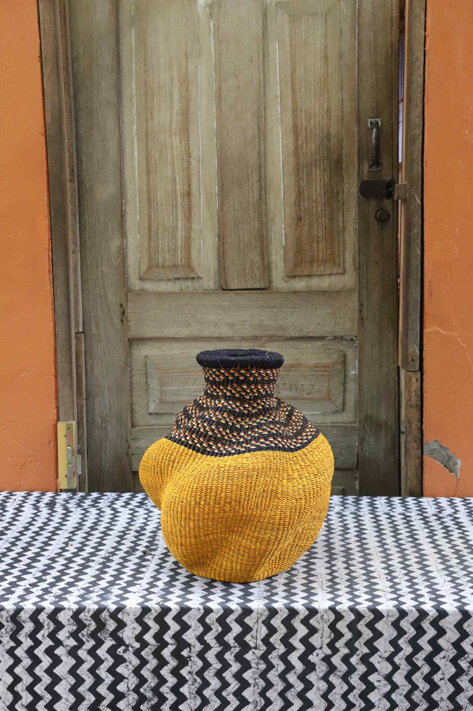 Handwoven Bolga art basket in an urn shape, dyed mustard yellow with a dark zigzag shoulder band.
