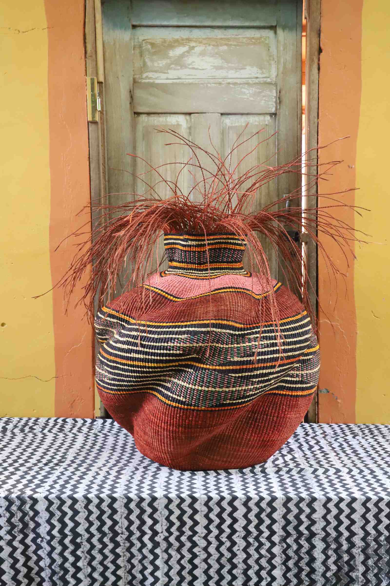 Round Bolga basket in rust red with a black and orange striped band, its narrow neck topped with loose straw fronds.