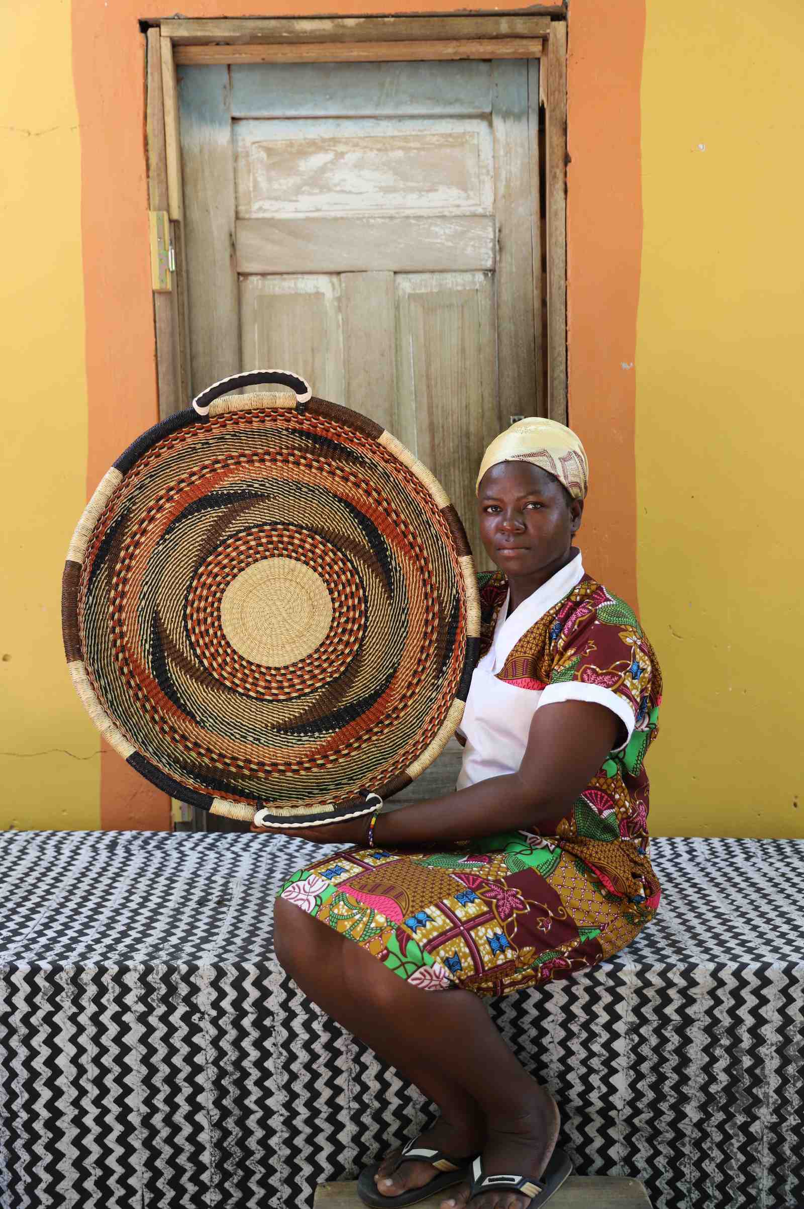 Medium handwoven Bolga wall tray disc with brown and orange bands, held by a seated woman in a printed dress.
