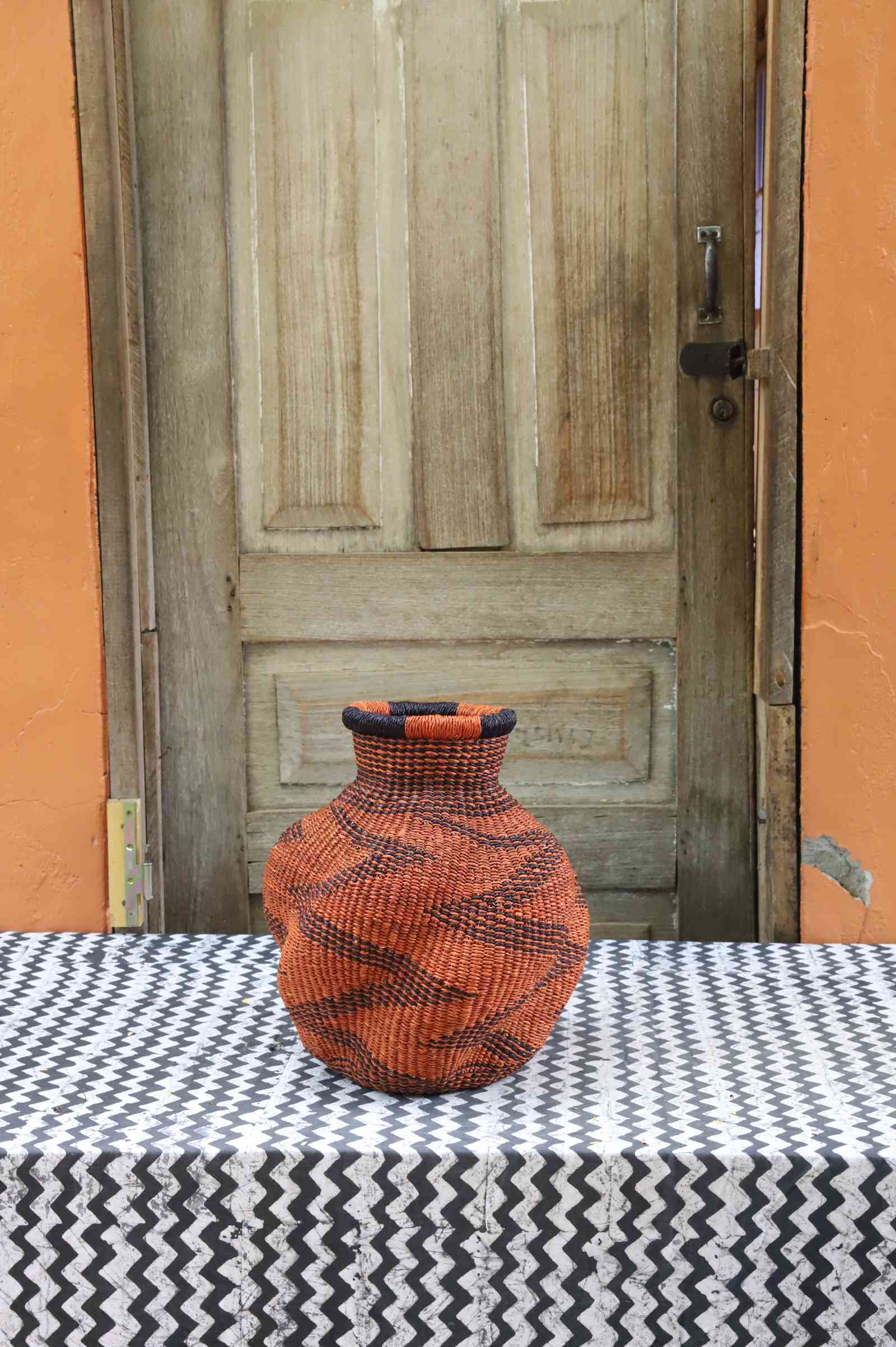 Handwoven Bolga art basket in an urn shape, dyed rust-orange with a dark zigzag wave motif and rim.