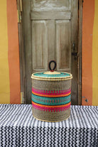 Handwoven Bolga laundry basket with coiled rim and lid in natural straw with pink, orange, green and teal fine stripes.