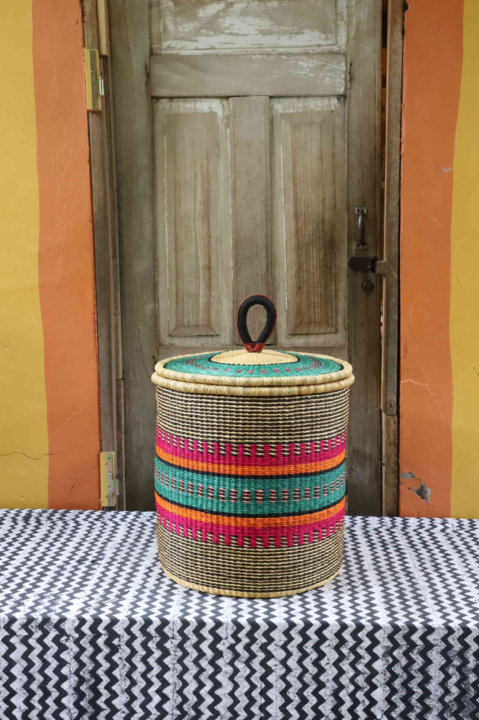 Handwoven Bolga laundry basket with coiled rim and lid in natural straw with pink, orange, green and teal fine stripes.