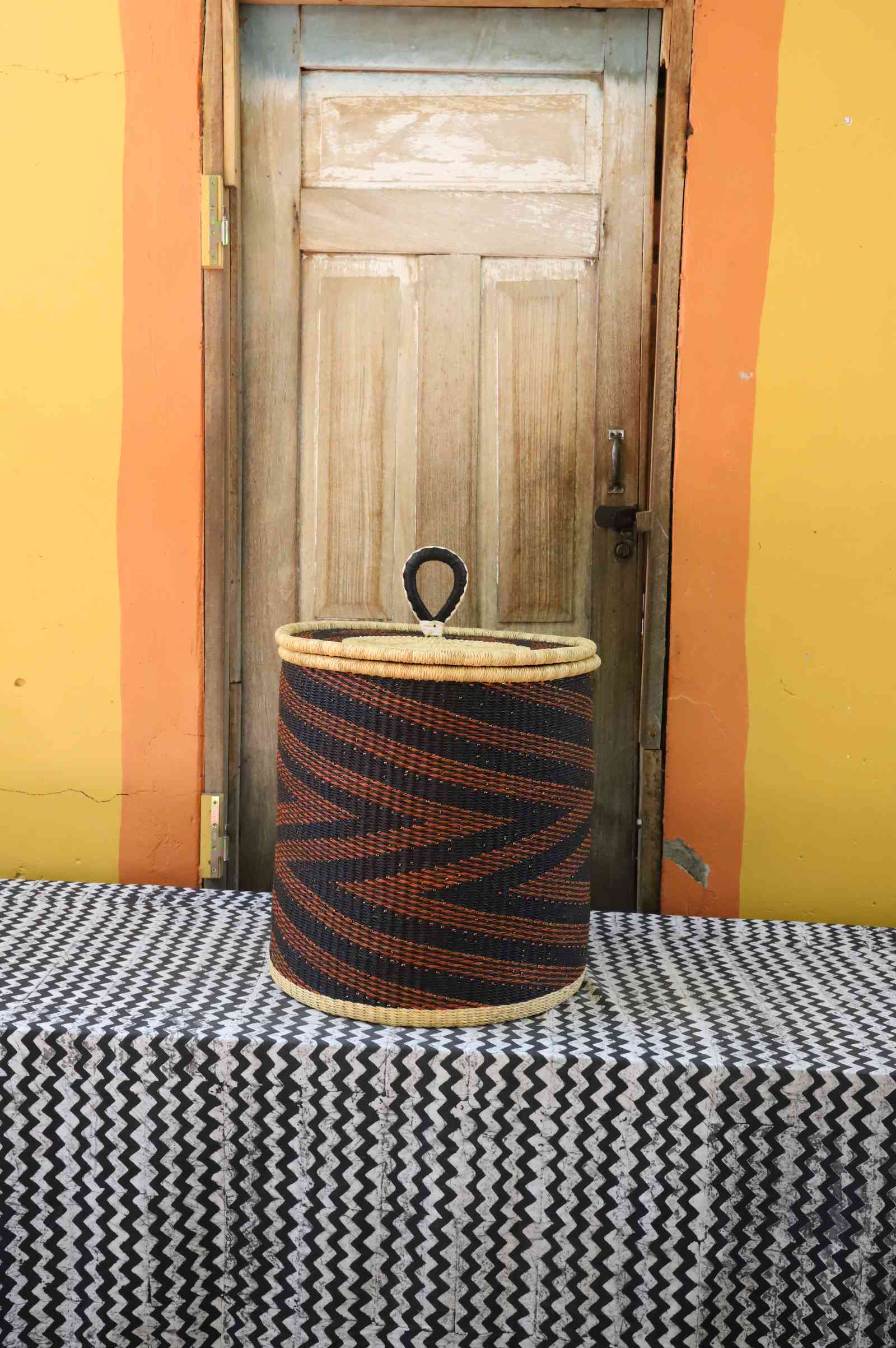Handwoven Bolga laundry basket with coiled rim and lid in dark navy with rust-orange diagonal wave stripes.