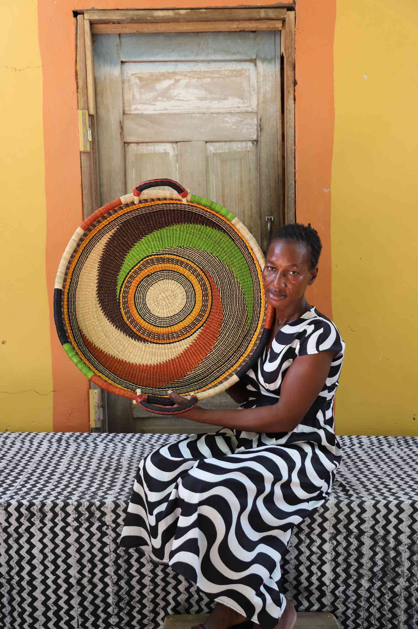 Medium handwoven Bolga wall tray disc with green and orange swirl, held by a seated woman in stripes.