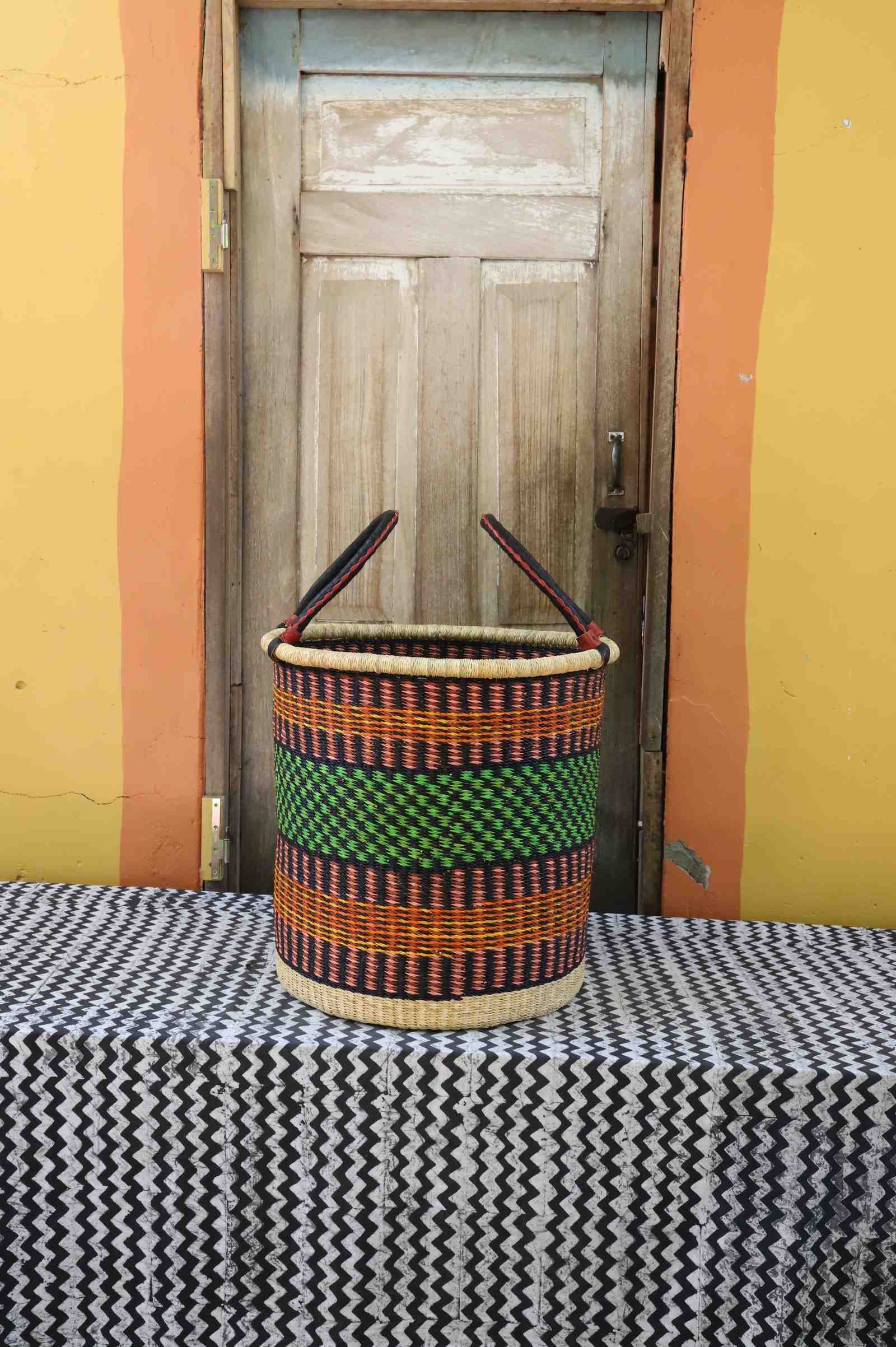 Large handwoven open-top Bolga laundry basket in dark straw with a green central band and orange stripes.