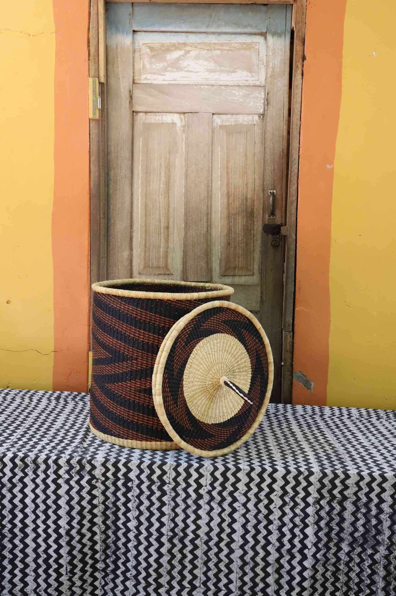 Handwoven Bolga laundry basket with lid removed, dark navy body with rust-orange waves and natural lid.