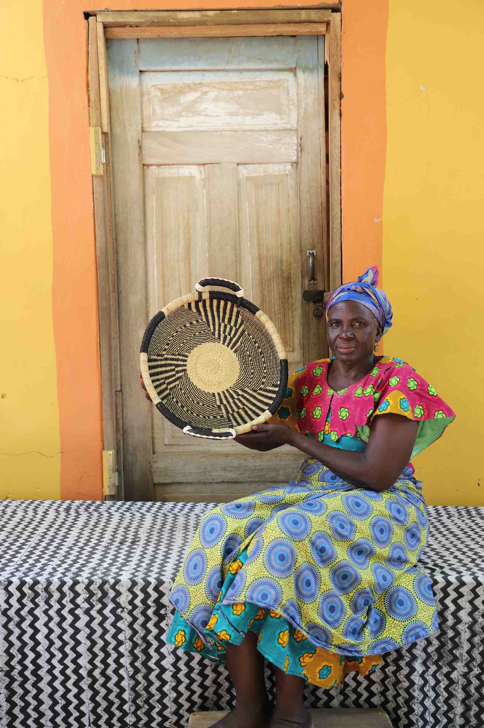 Handwoven Bolga wall tray disc in dark straw with fine radial stripes, held by a seated woman in patterned dress.